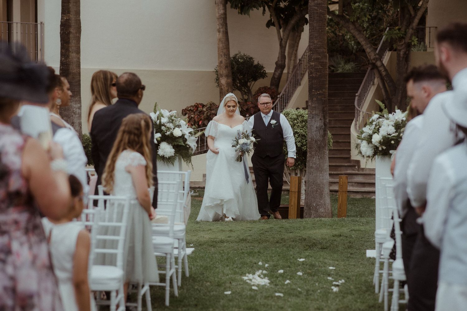 An outdoor wedding ceremony under tropical trees with white chairs and floral arrangements.Tenerife Wedding Photographer at Hotel Vincci