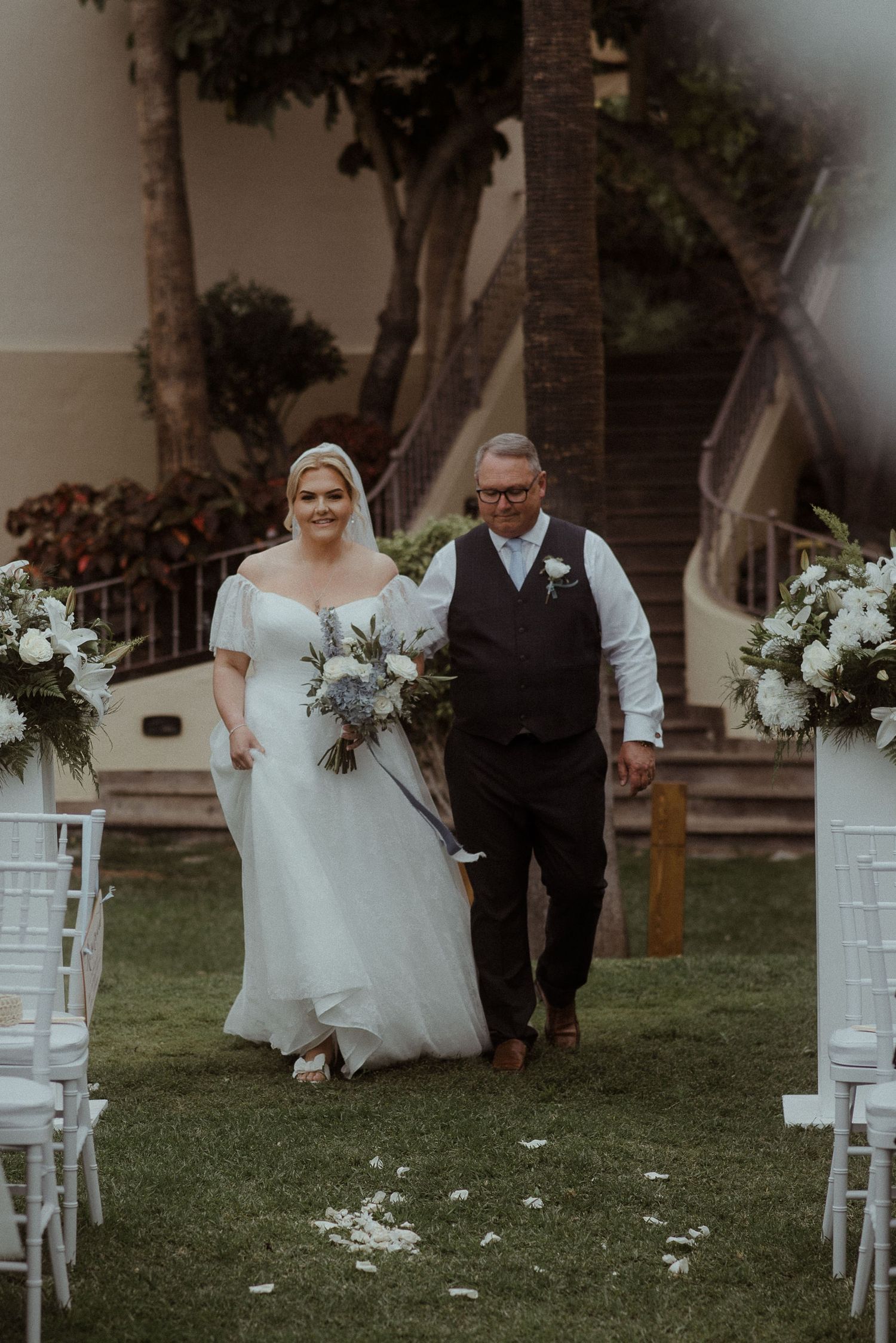 An outdoor wedding processional features white chairs and floral arrangements lining the aisle at a garden venue. Tenerife Wedding