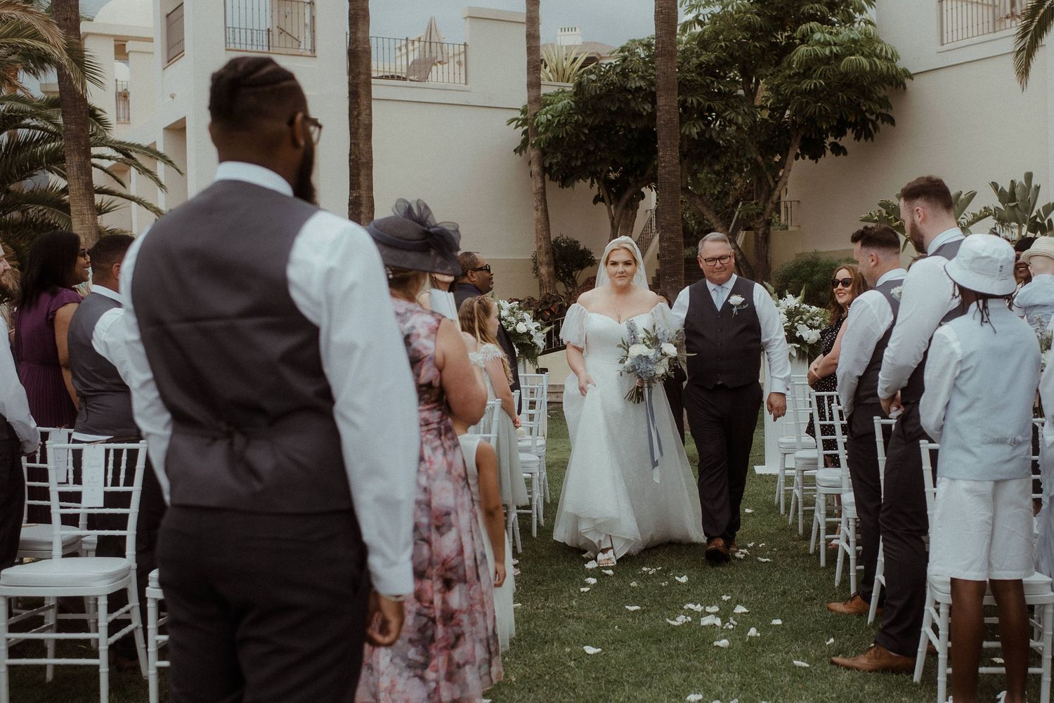 Wedding guests in formal attire stand along white chairs during an outdoor ceremony. Tenerife Wedding Photographer at Hotel Vincci