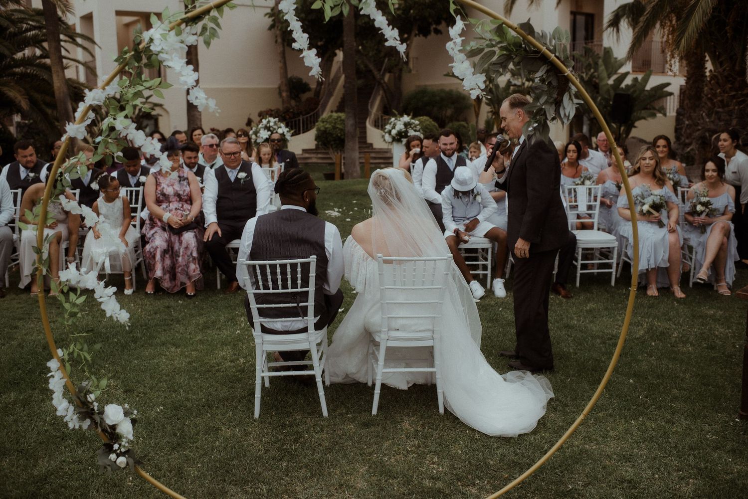 A wedding ceremony under a floral arch with guests seated on white chairs in a garden setting. Tenerife Wedding Photographer at Hotel Vincci