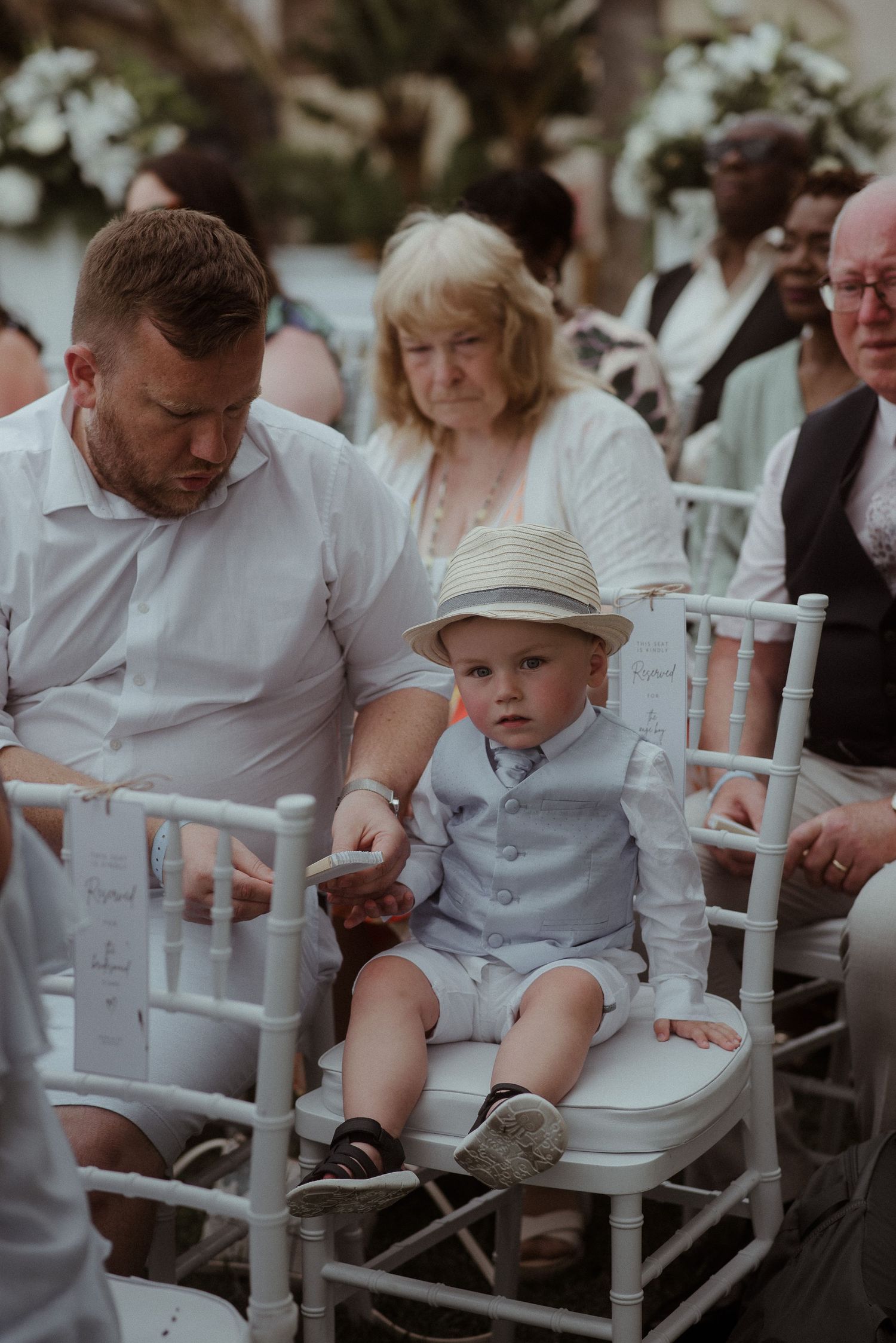 A young guest in formal attire sits attentively during an outdoor wedding ceremony. Tenerife Wedding Photographer at Hotel Vincci