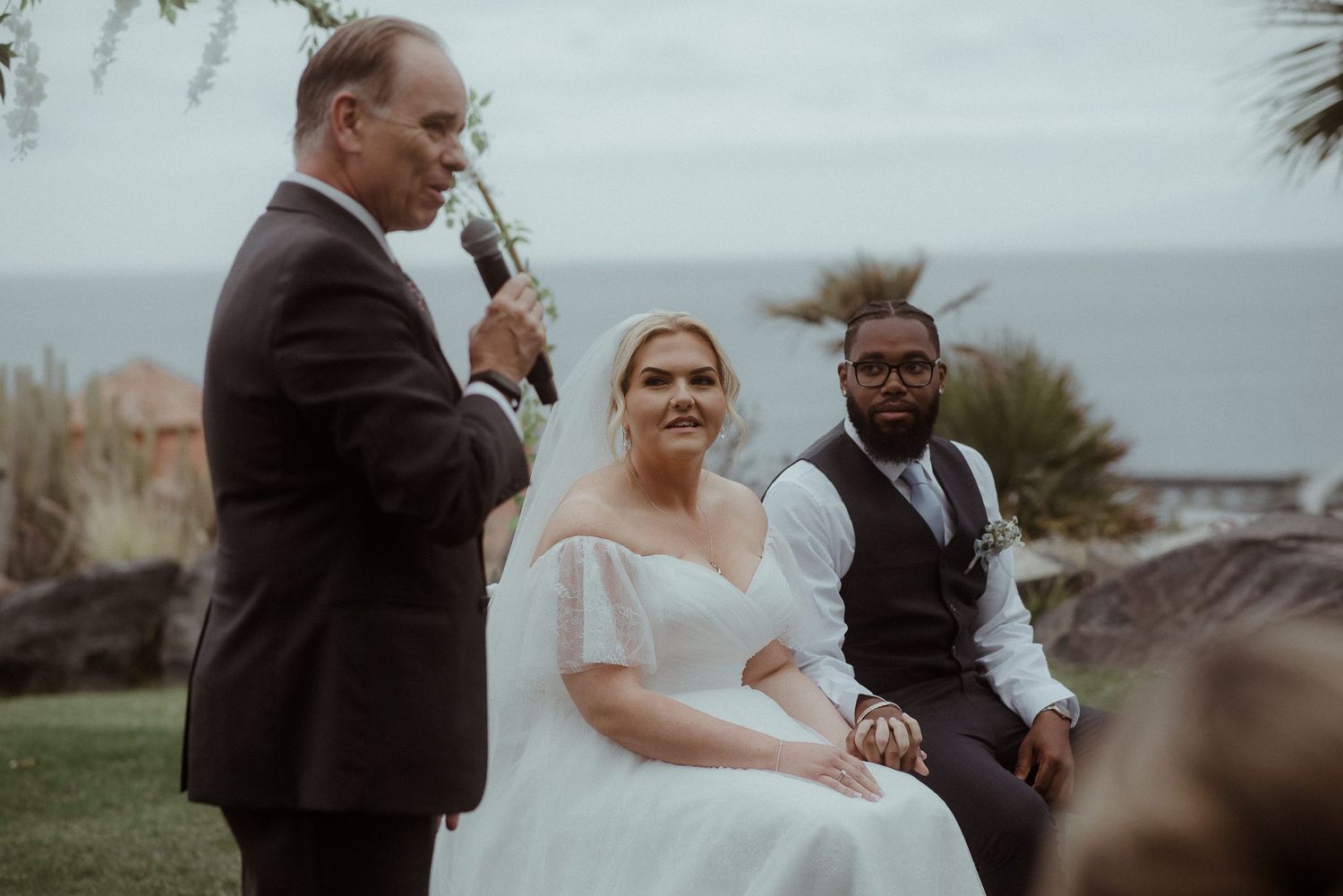 An outdoor wedding ceremony with palm trees and ocean views features speakers addressing the gathering. Tenerife Wedding Photographer