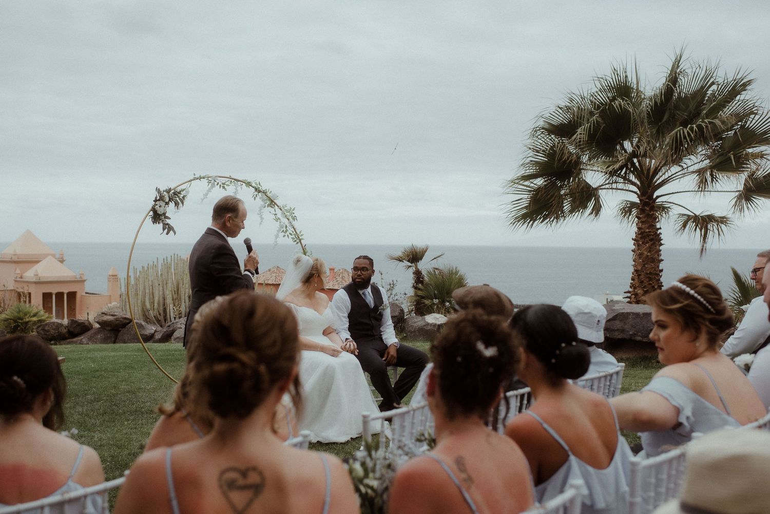 Wedding guests in blue dresses watch the ceremony under a floral arch with ocean views. Tenerife Wedding Photographer at Hotel Vincci