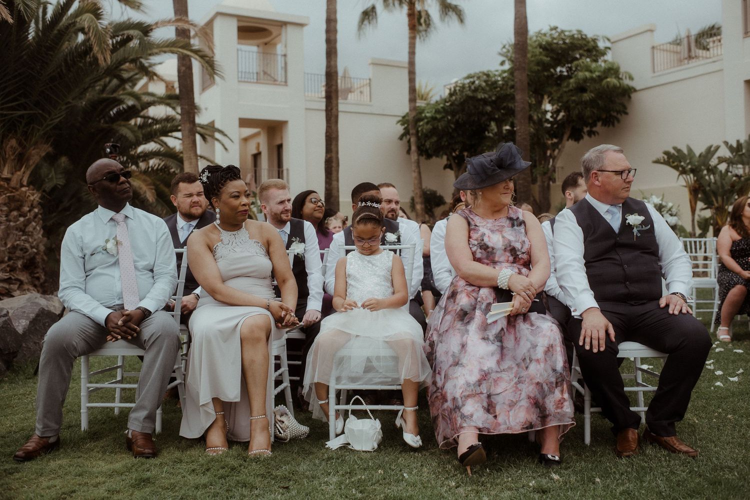 Wedding guests seated in white chairs on a lawn with palm trees in the background. Tenerife Wedding Photographer at Hotel Vincci