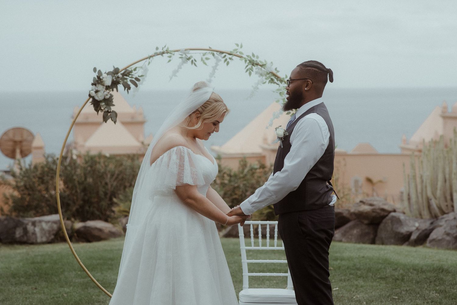 Wedding ceremony under a circular floral arch with ocean and buildings in background. Tenerife Wedding Photographer at Hotel Vincci