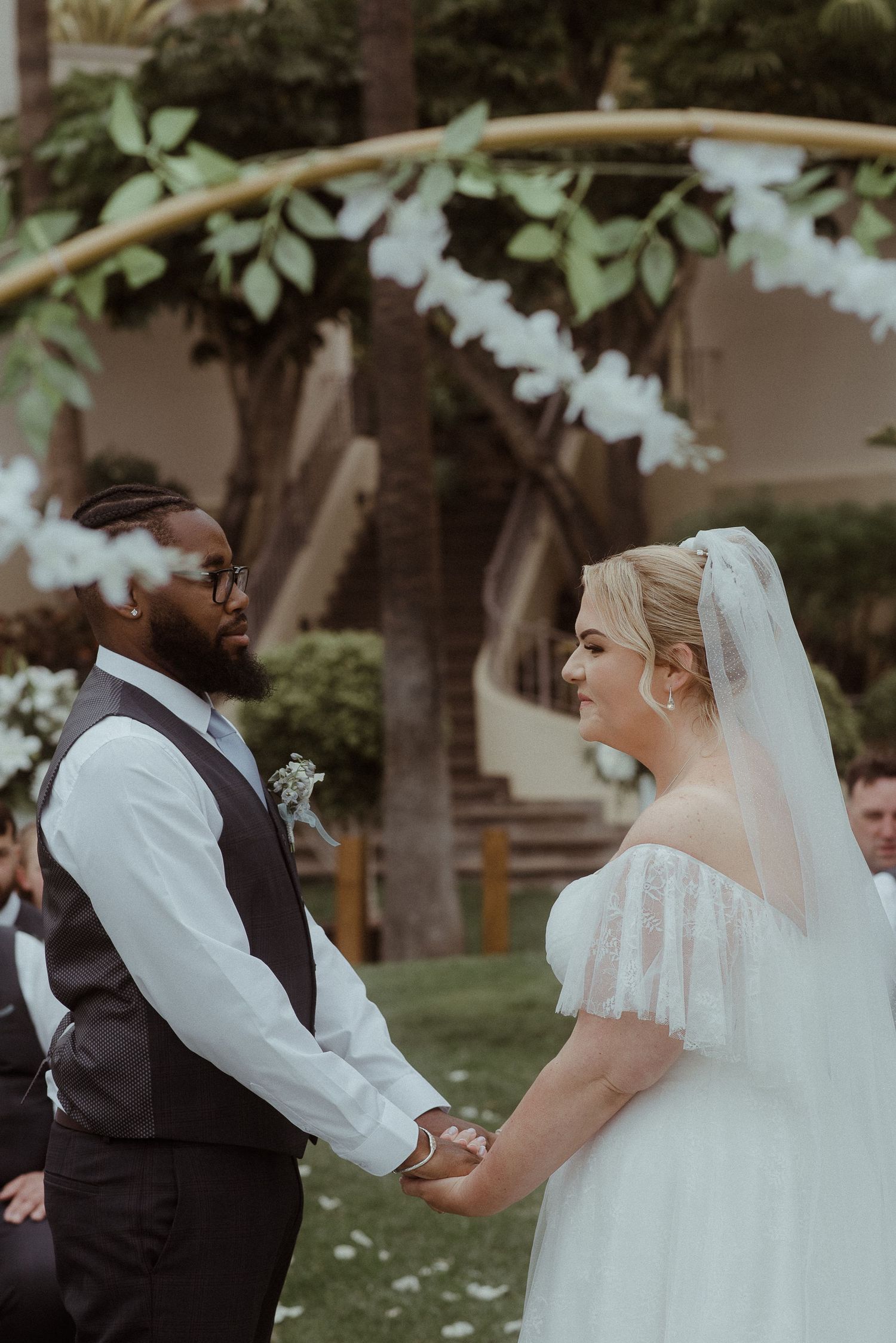 Couple holds hands during outdoor wedding ceremony under floral archway.
