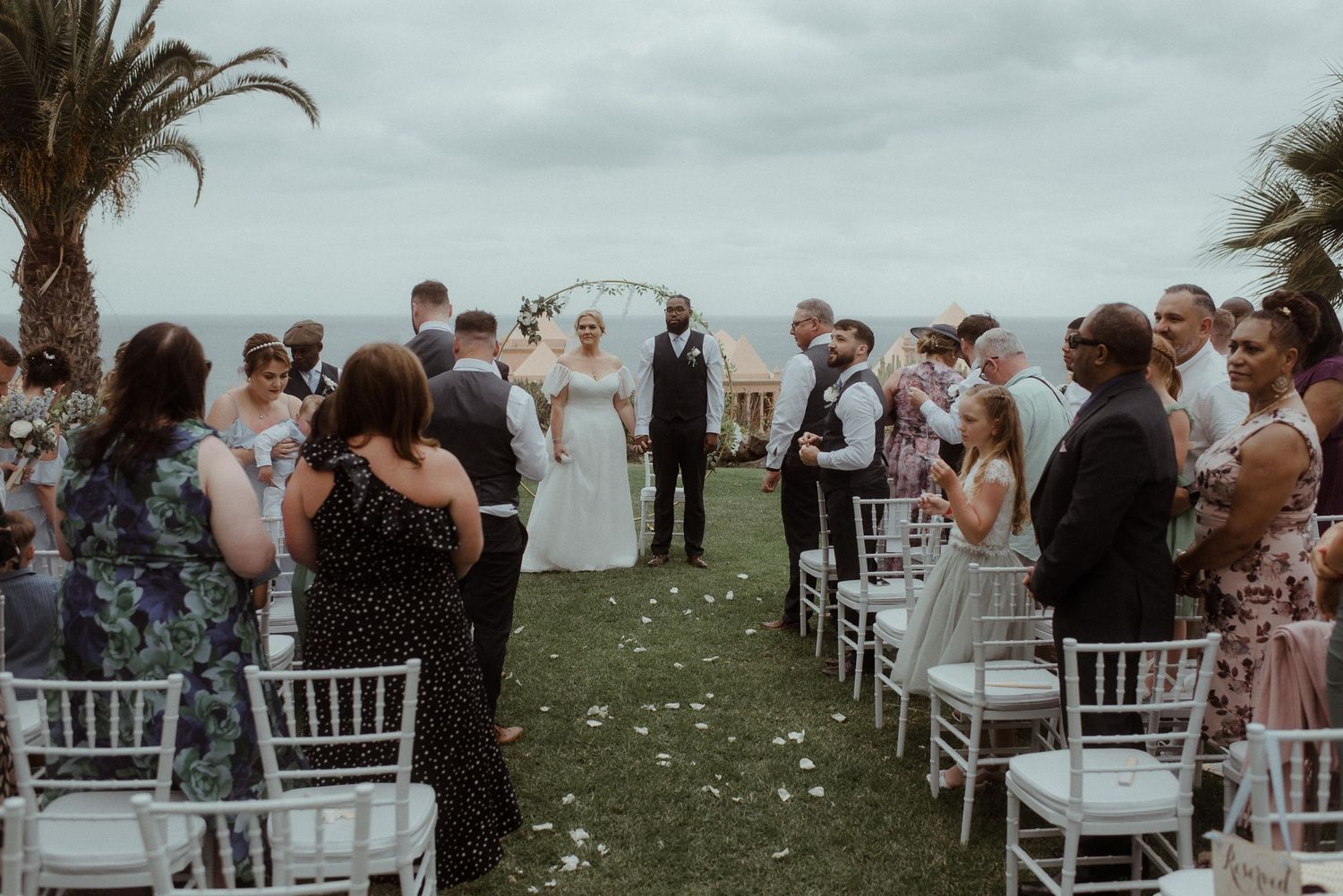 Wedding guests stand at a seaside ceremony with white chairs and palm trees under moody skies.