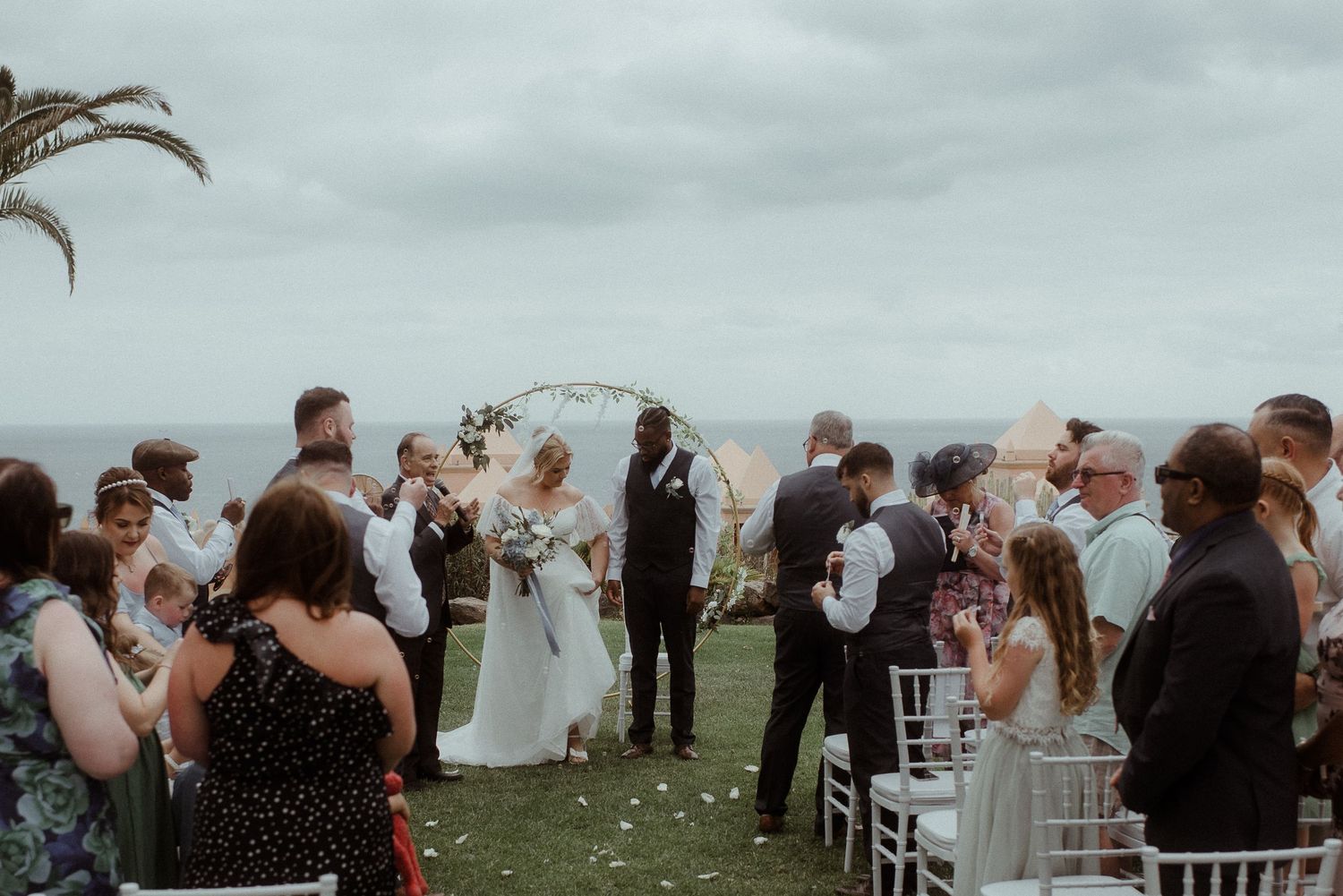 Outdoor wedding ceremony with white chairs and palm trees against a cloudy coastal backdrop.