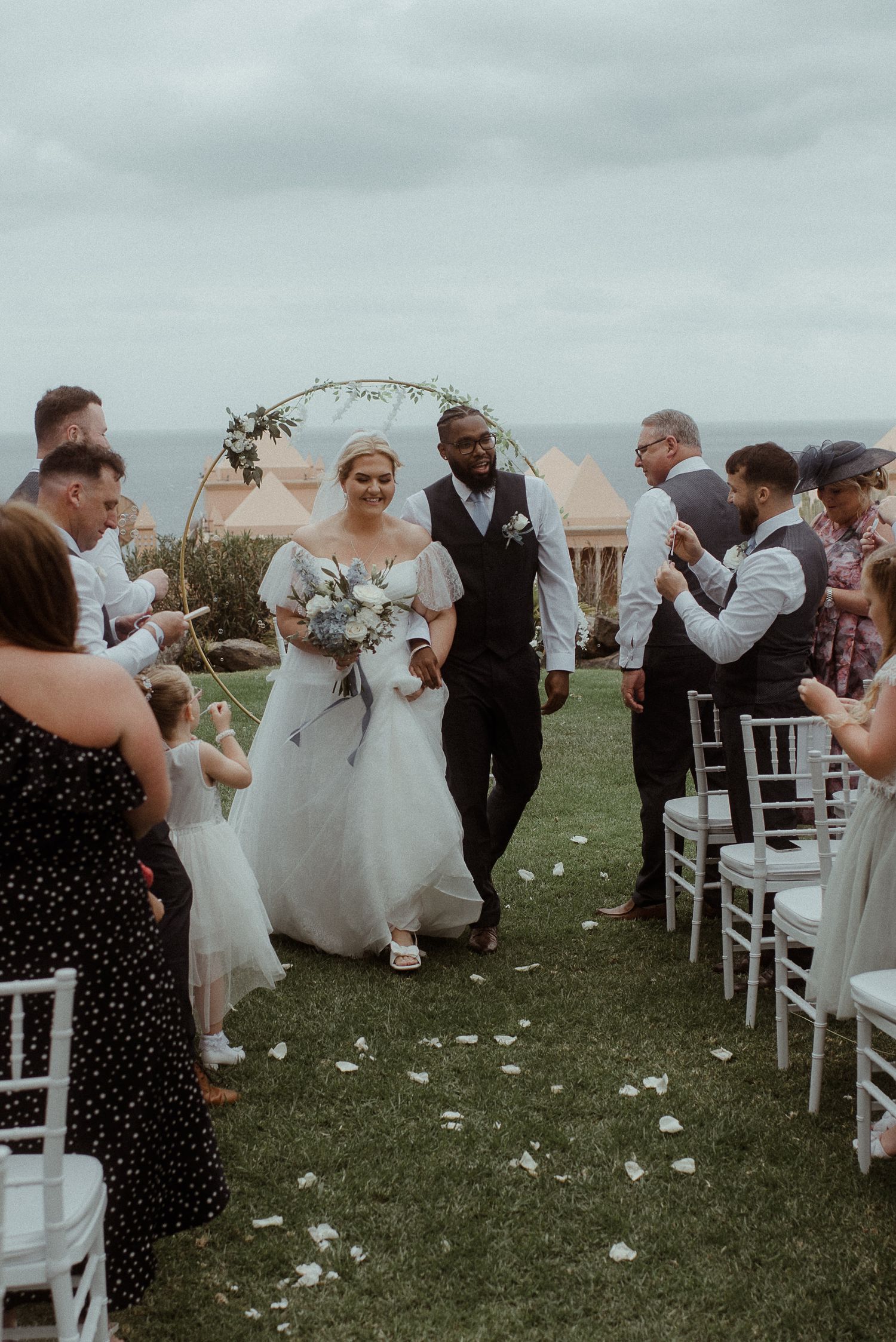 Newlyweds walk down the aisle at outdoor beach wedding with floral arch and white chairs.