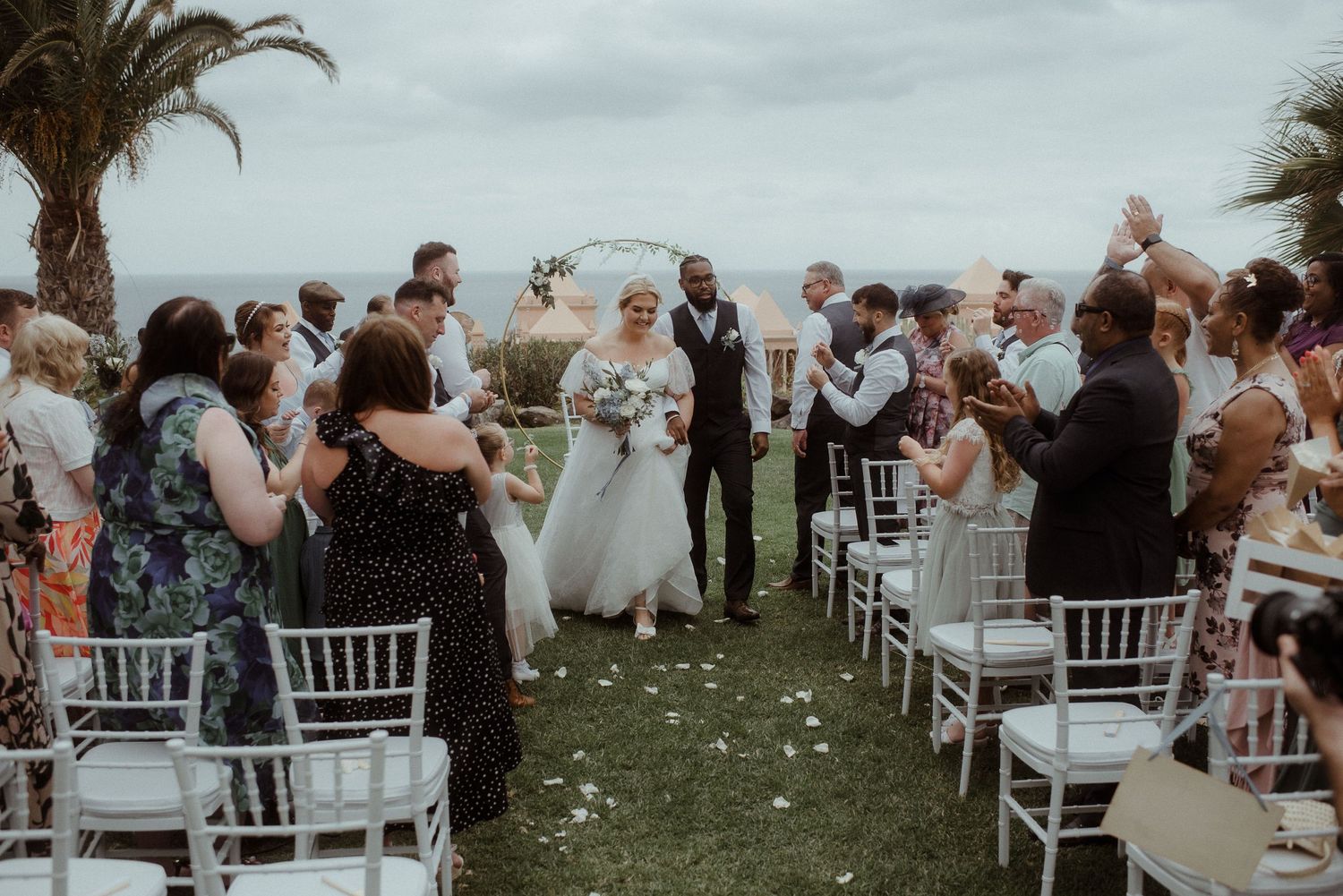 Guests celebrate at coastal wedding ceremony with palm trees and scattered flower petals.