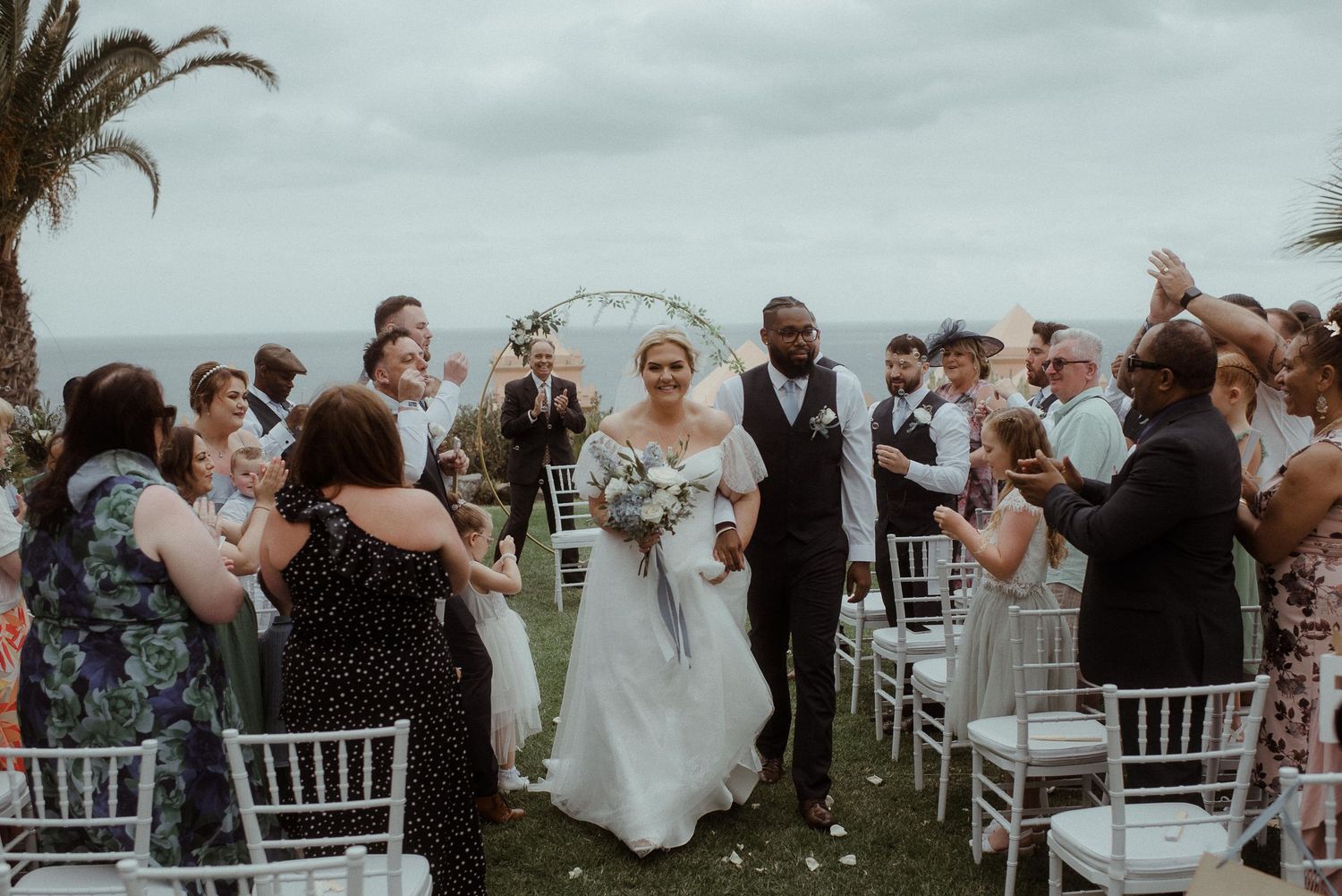 Wedding guests cheer during recessional at outdoor ceremony with white chairs and floral decor.