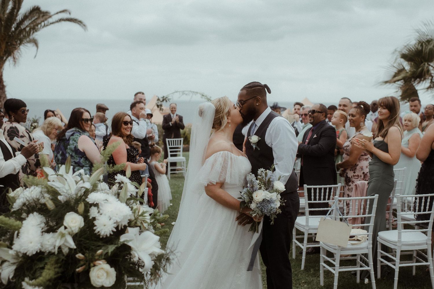 Coastal wedding ceremony with white floral arrangements and palm trees under cloudy skies.
