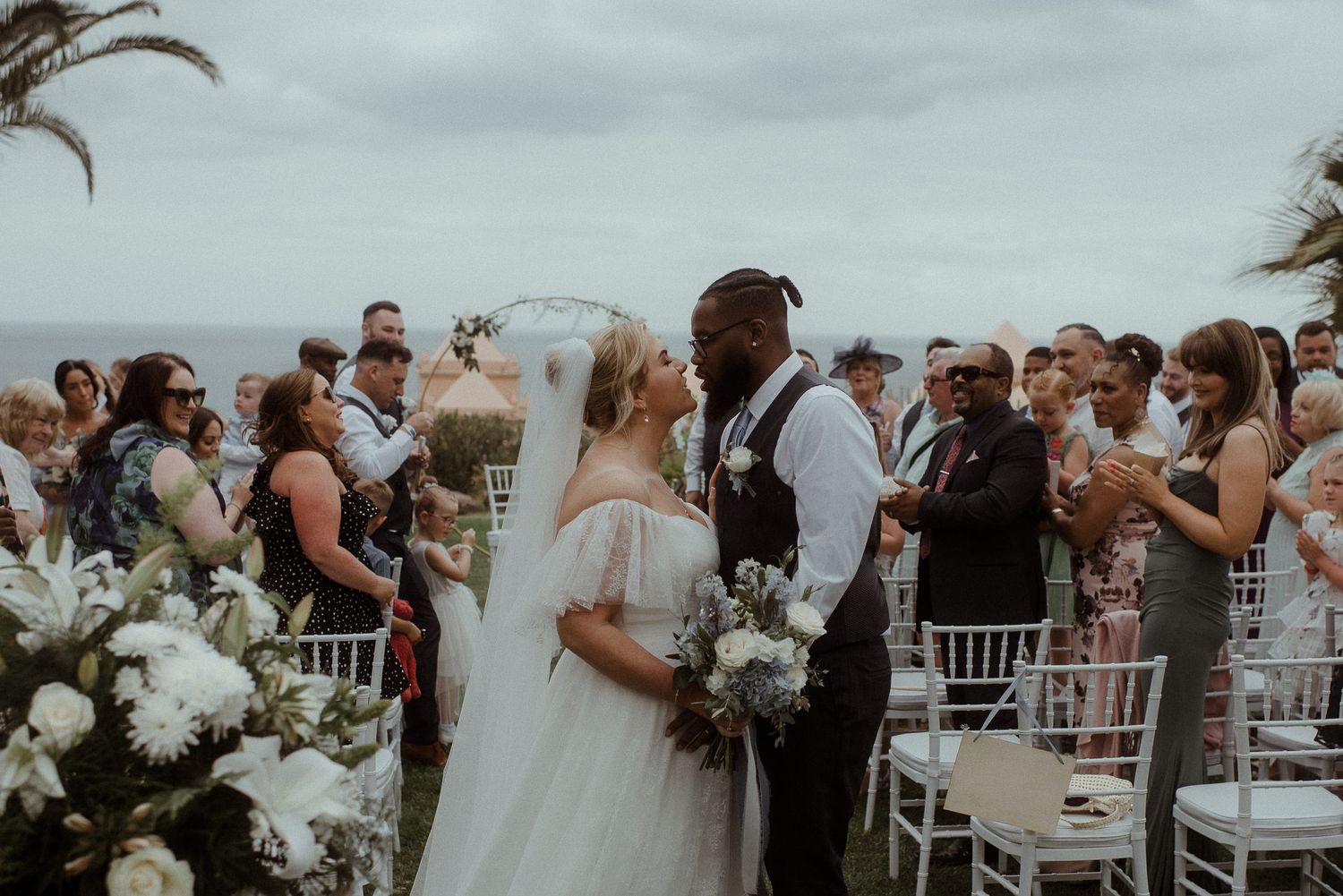 Romantic outdoor wedding ceremony with white chairs and tropical palm trees against grey sky.
