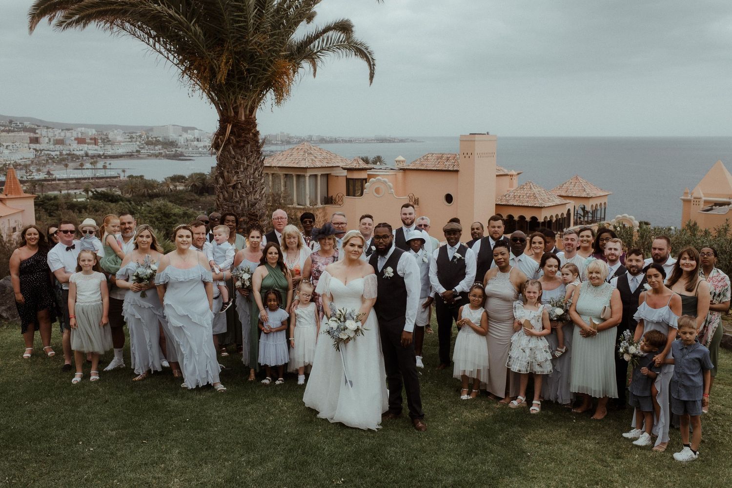 Large wedding group photo with palm trees and ocean views in the background.