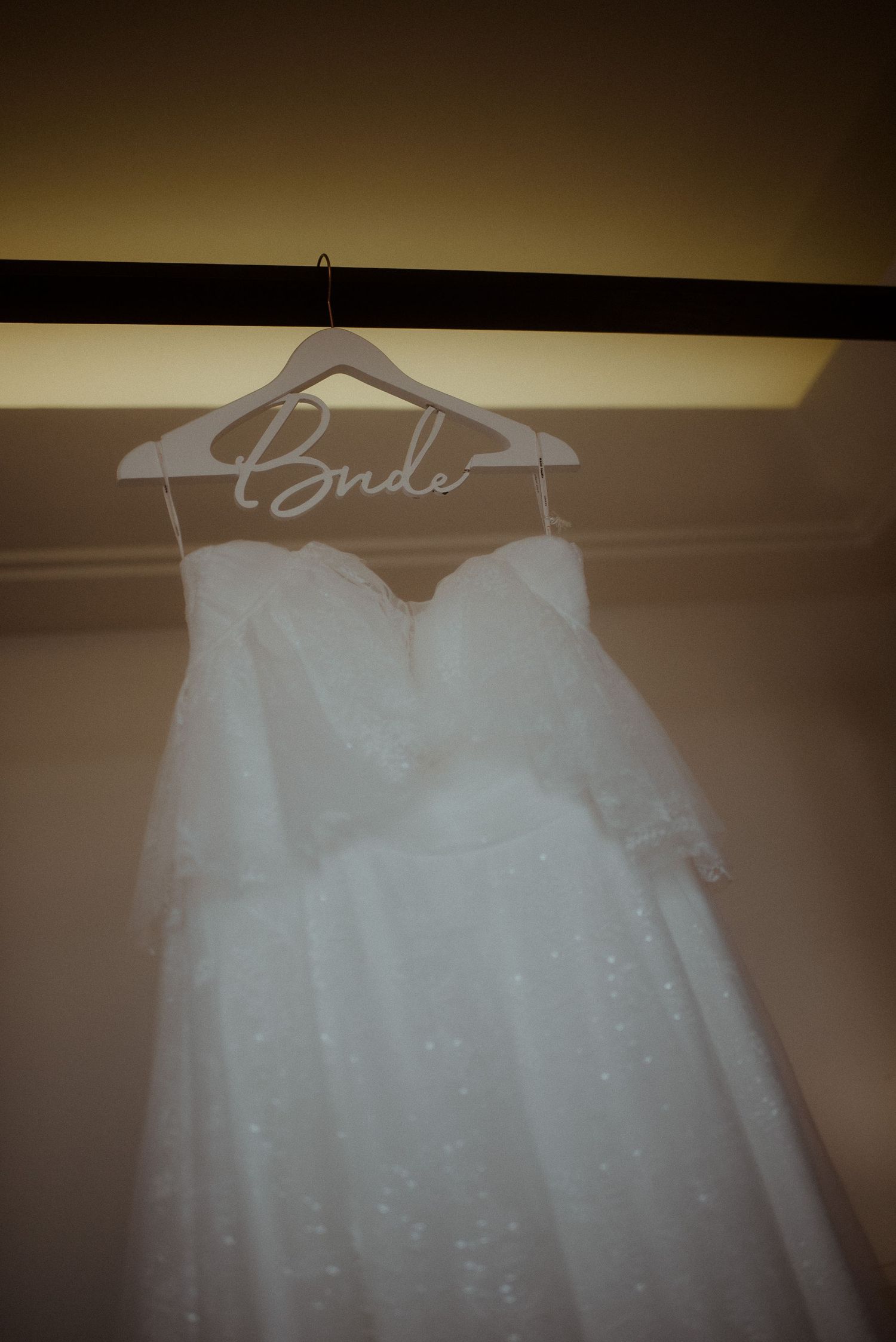 A white wedding dress with sparkly details hanging on a custom 'Bride' hanger against a neutral wall.