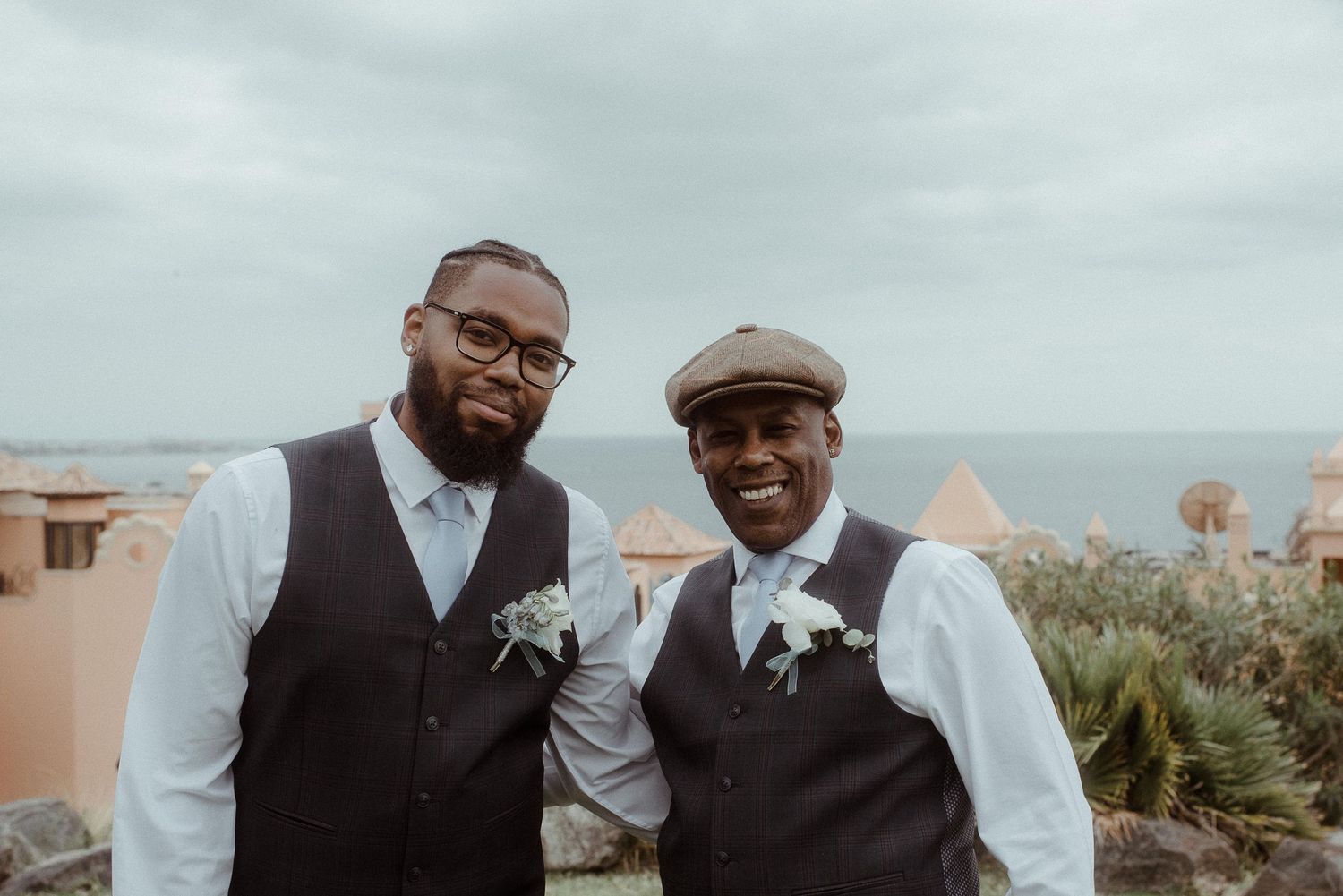 Two groomsmen in matching vests pose for a photo at a coastal venue.