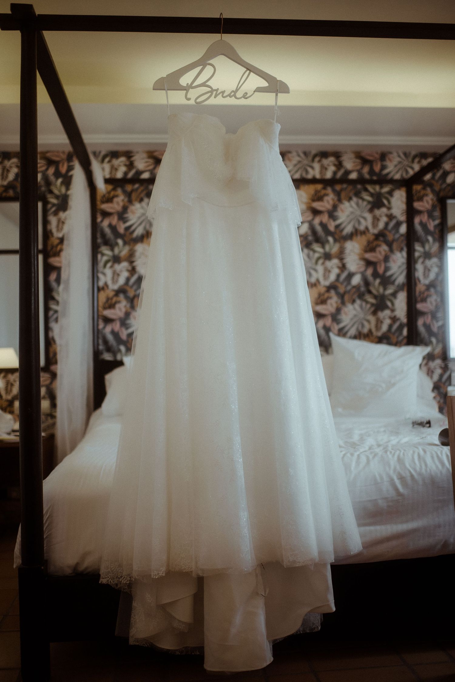 Elegant white wedding gown suspended from a 'Bride' hanger in a bedroom with floral wallpaper. Tenerife Wedding Photographer at Hotel Vincci