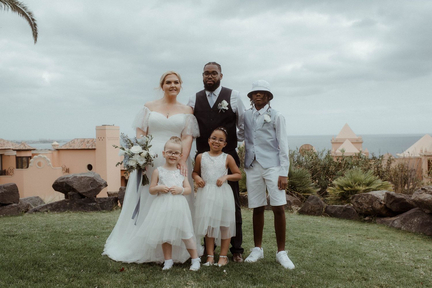 Wedding party members pose for a formal photo on a lawn with Mediterranean architecture in background.