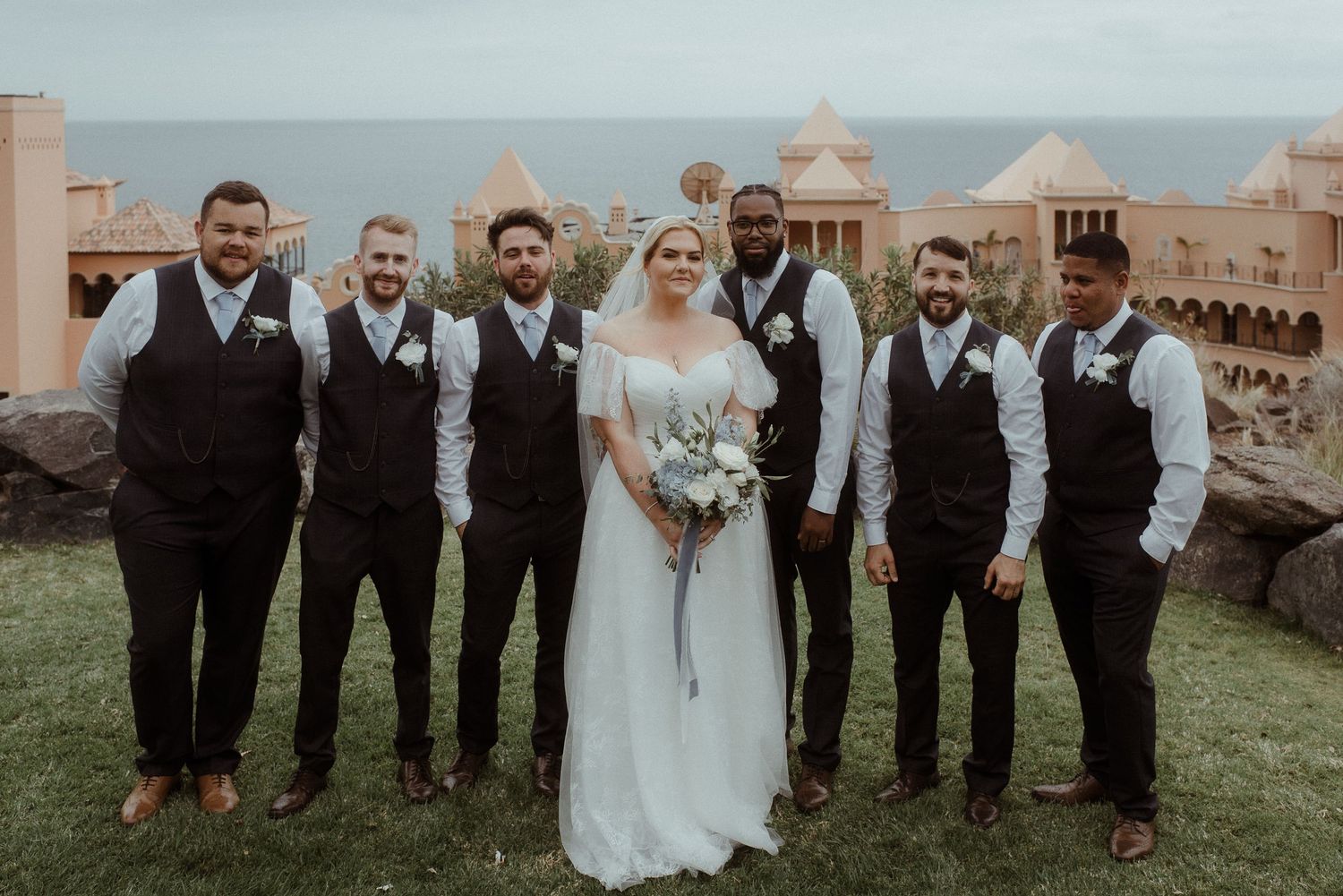 Groomsmen in matching black vests pose with couple at Mediterranean-style coastal venue.