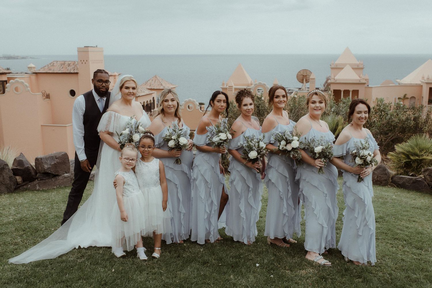Bridesmaids in pale blue dresses pose with bride holding bouquets against ocean backdrop.