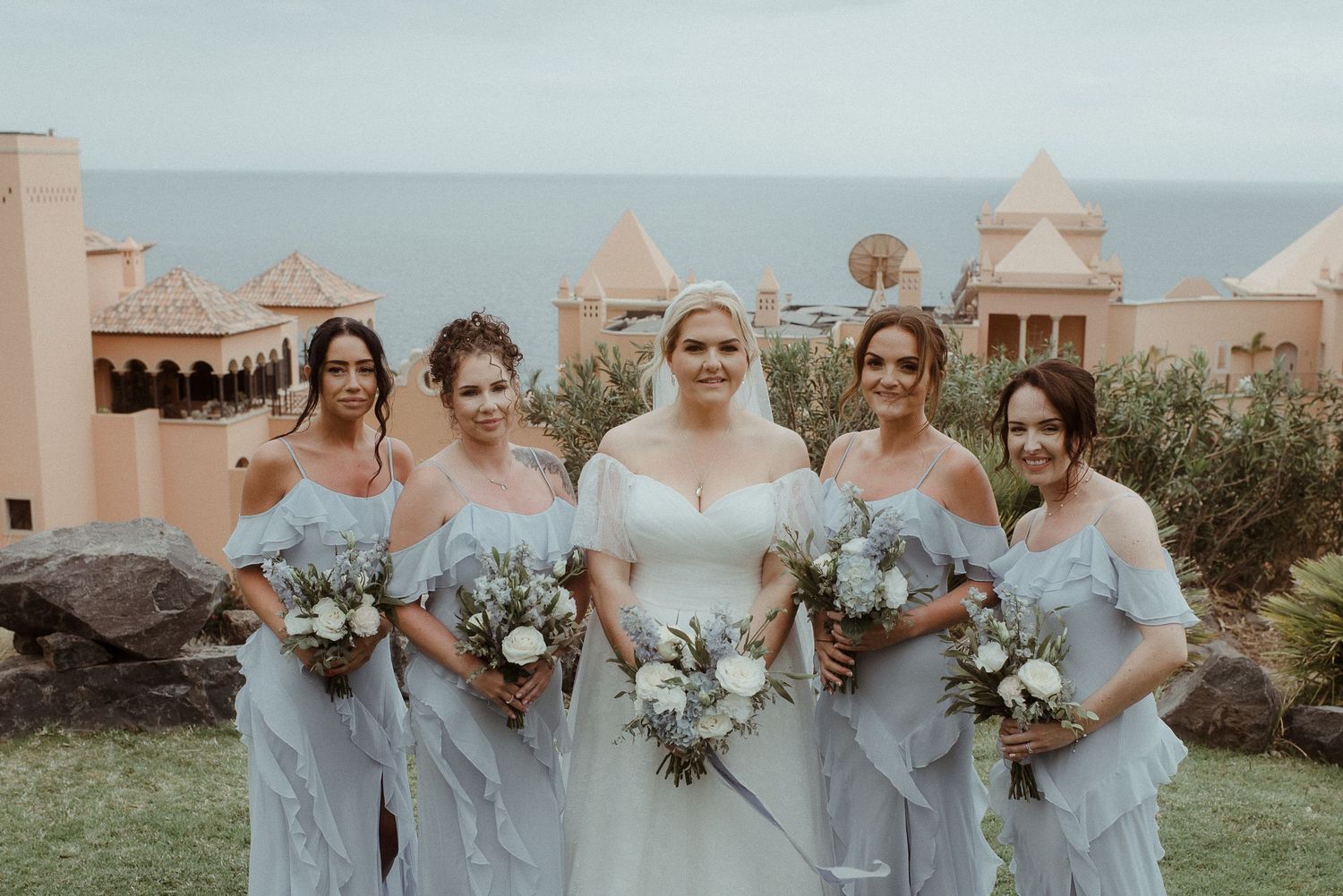 Bridal party in soft blue dresses holding white floral bouquets pose on coastal lawn.