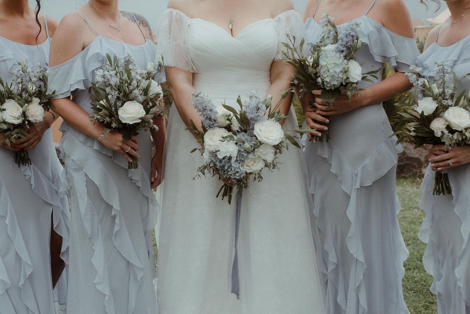 Bridesmaids in flowing gray dresses hold elegant white and green floral bouquets against an outdoor backdrop.