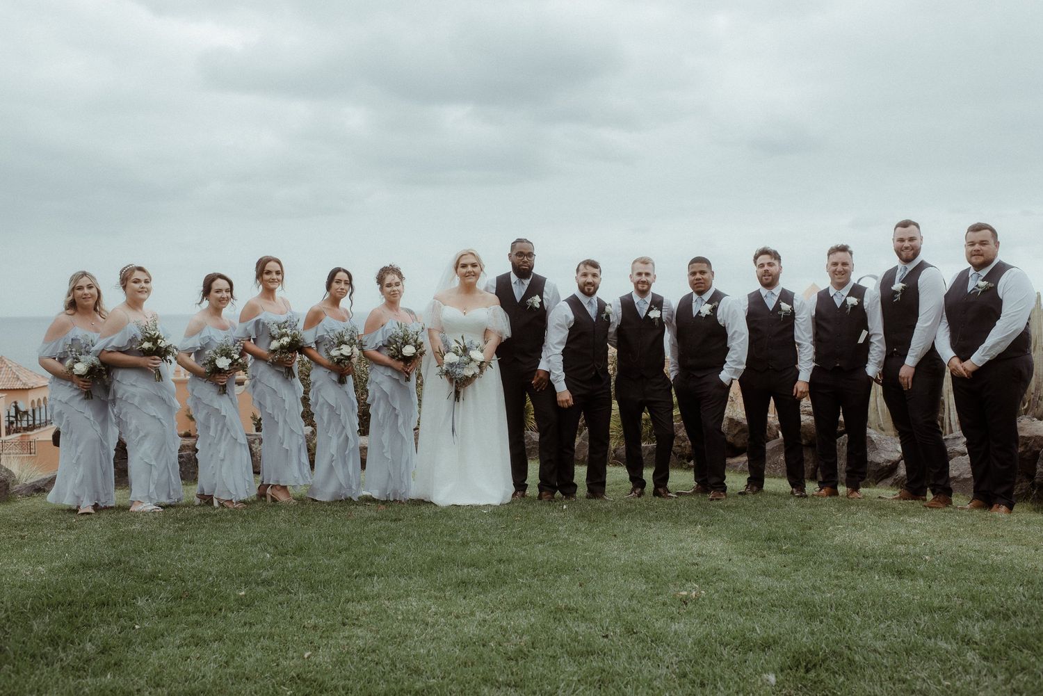 Wedding party poses on grassy hillside with ocean view in gray and black formal attire.