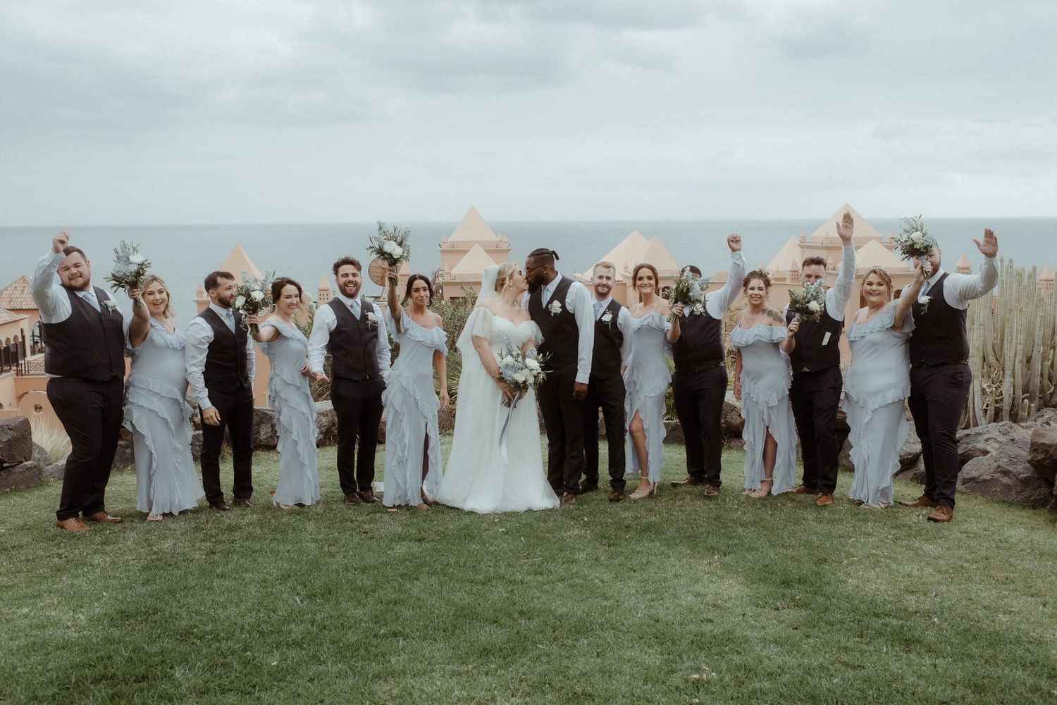 Wedding party celebrates with raised arms on scenic coastal overlook.