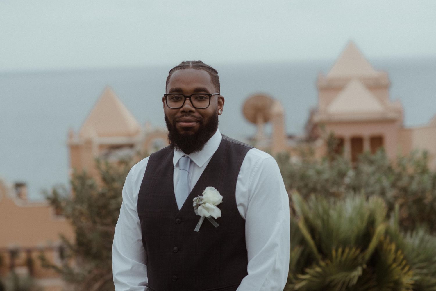 Wedding attendant in formal vest stands before terra cotta resort buildings and ocean view.