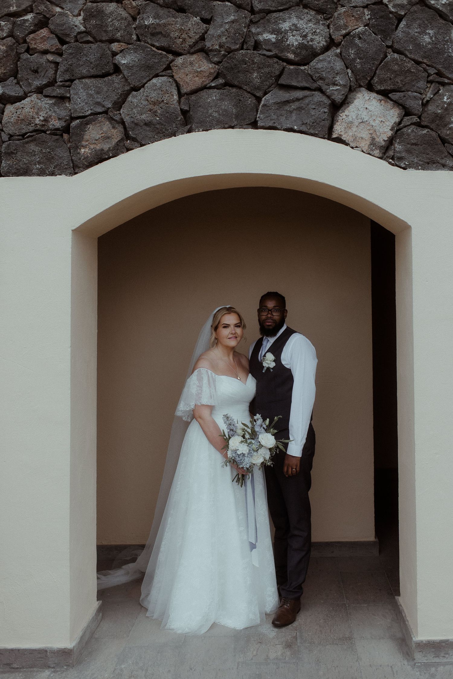 Elegant wedding portrait of couple standing in stone archway wearing formal attire.