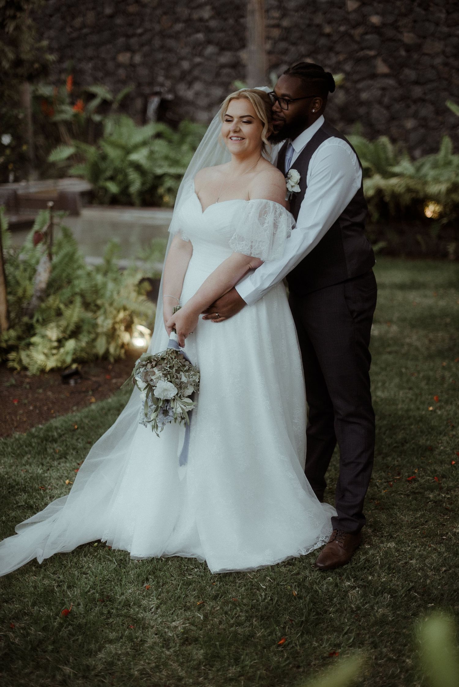 A romantic wedding portrait of newlyweds embracing in a garden setting with greenery in the background.
