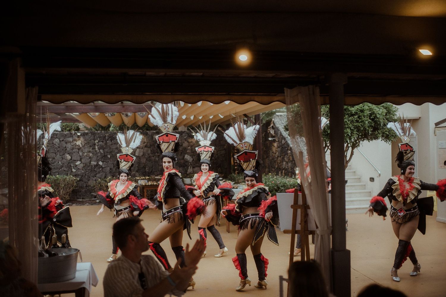 Traditional performers in feathered headdresses and matching outfits dance under covered outdoor venue.