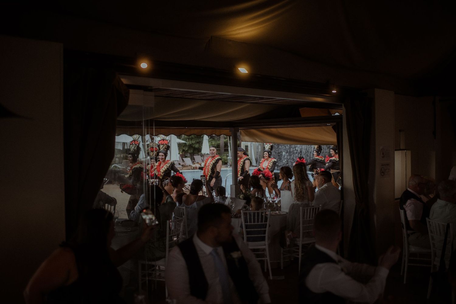 Wedding reception guests watch entertainers perform through a doorway in dimly lit venue.