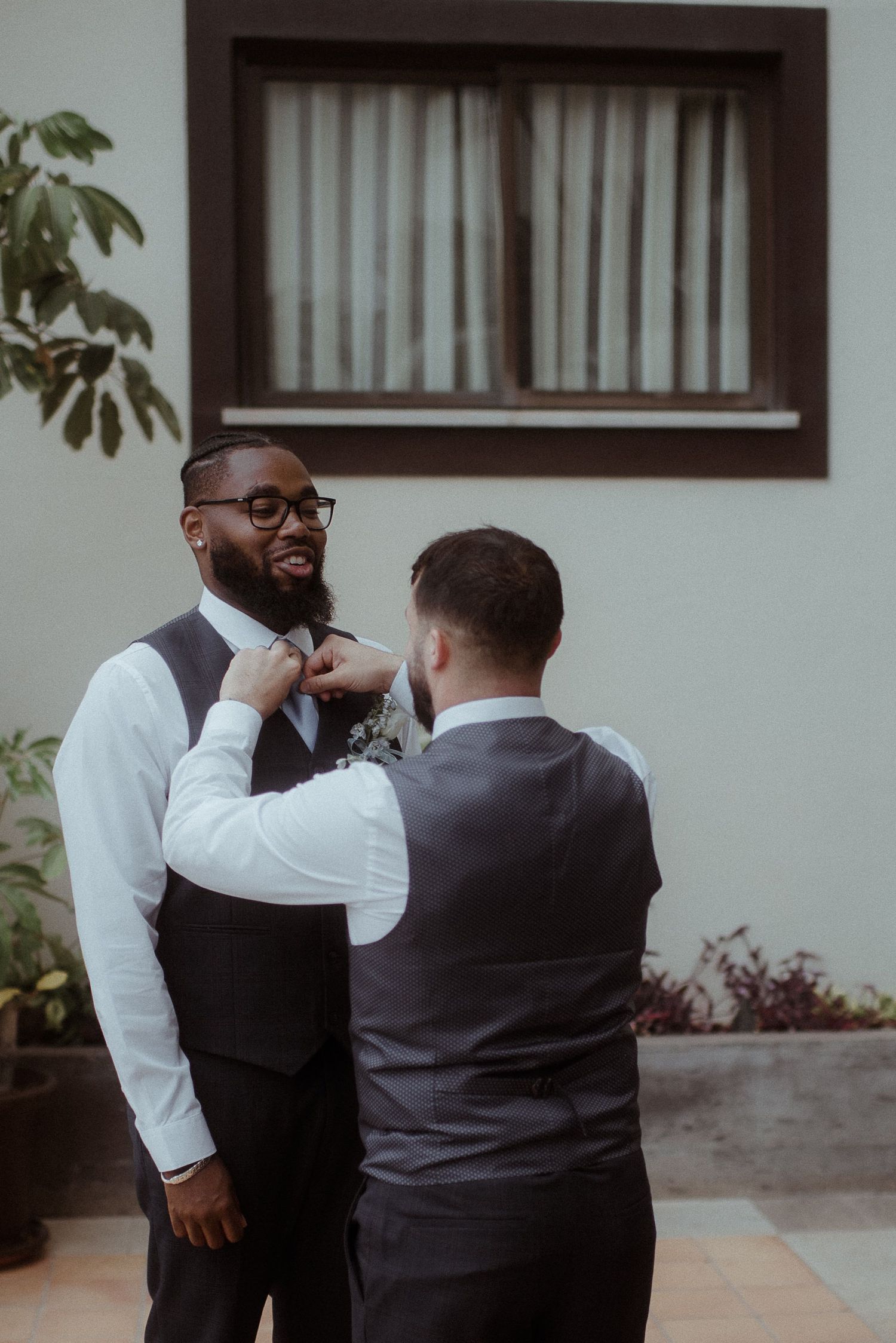 Two people in formal vests and white shirts adjusting a tie before an event.