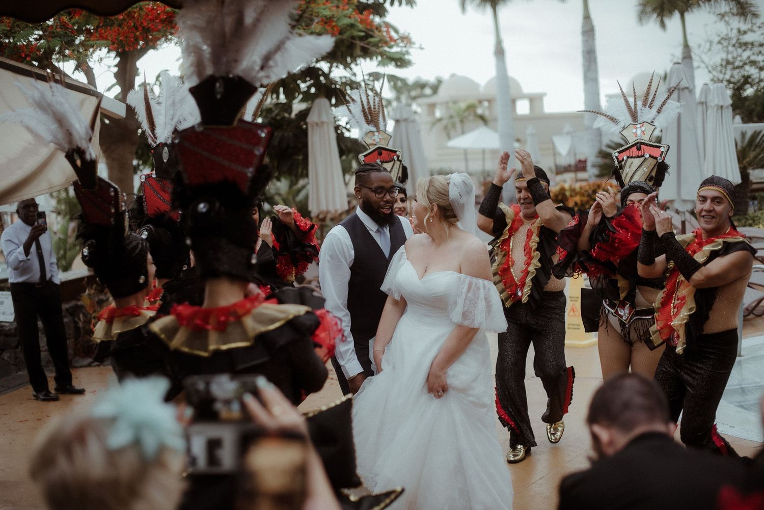 Outdoor wedding reception with costumed performers dancing amid palm trees and guests.