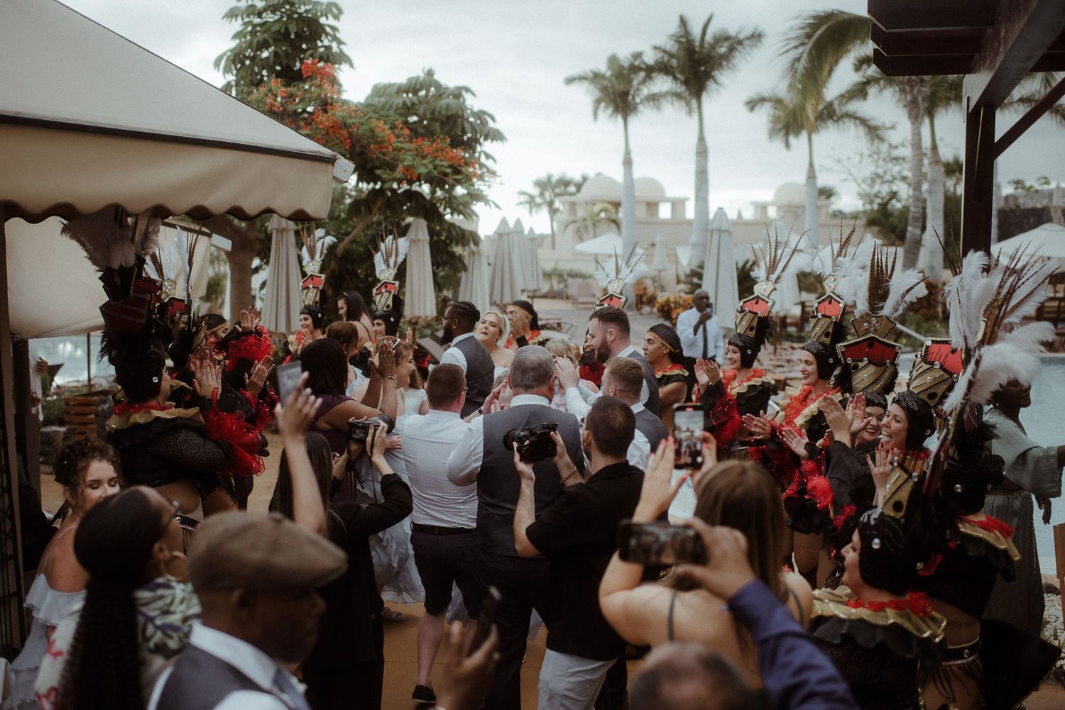 Crowded outdoor wedding reception with performers and guests dancing under palm trees.