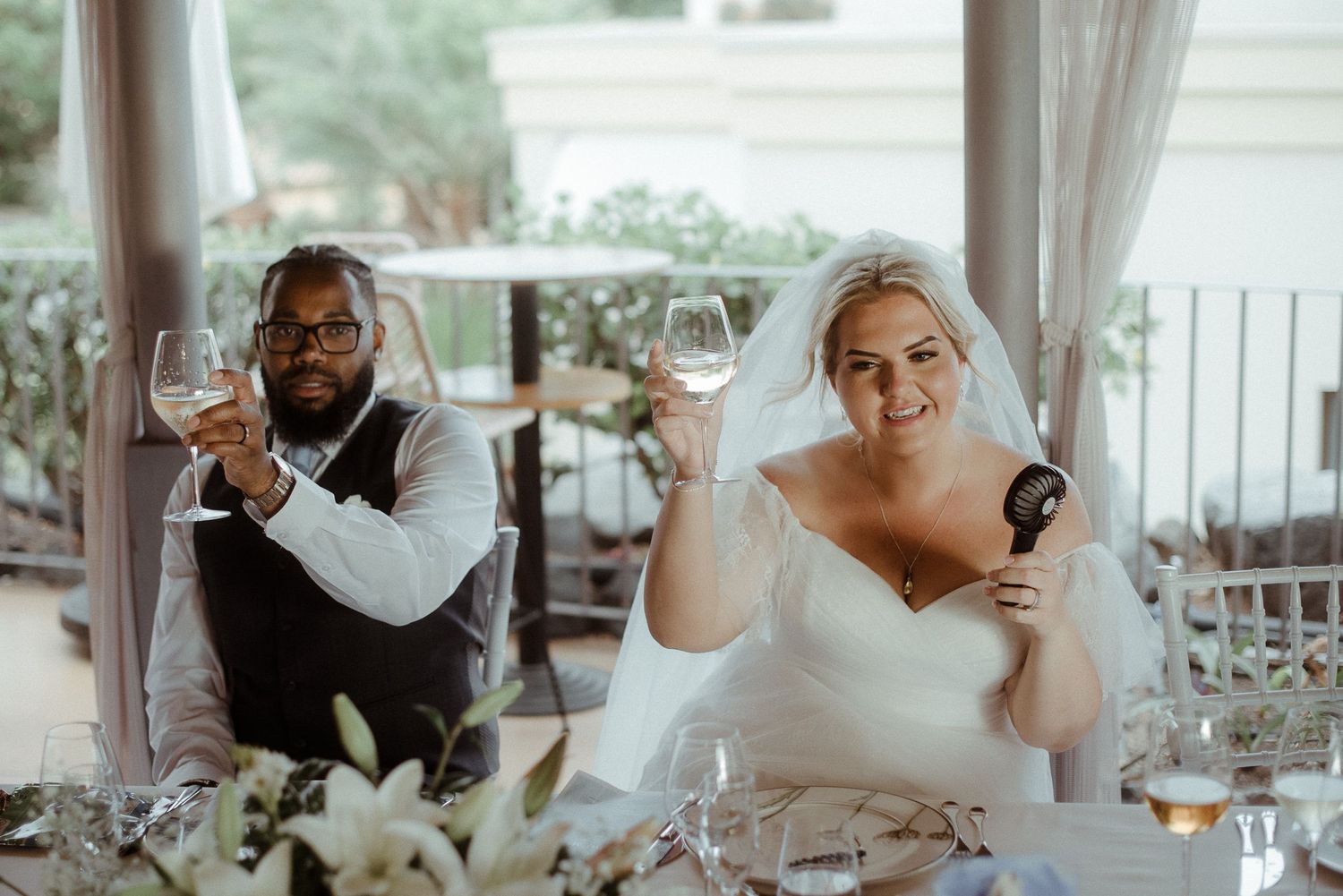 A couple toasts with wine glasses at their wedding reception table.