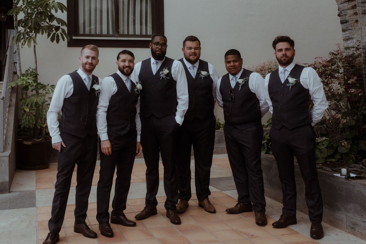 Wedding party of six groomsmen in matching dark vests and white shirts posing together.