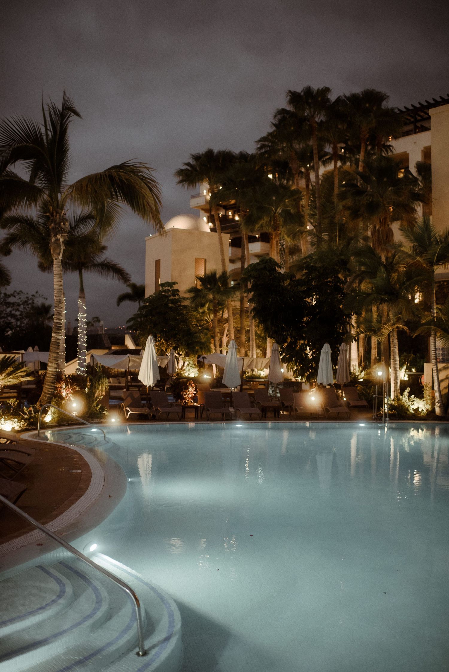 Illuminated resort pool at night with palm trees and white cabanas creating a tropical ambiance.