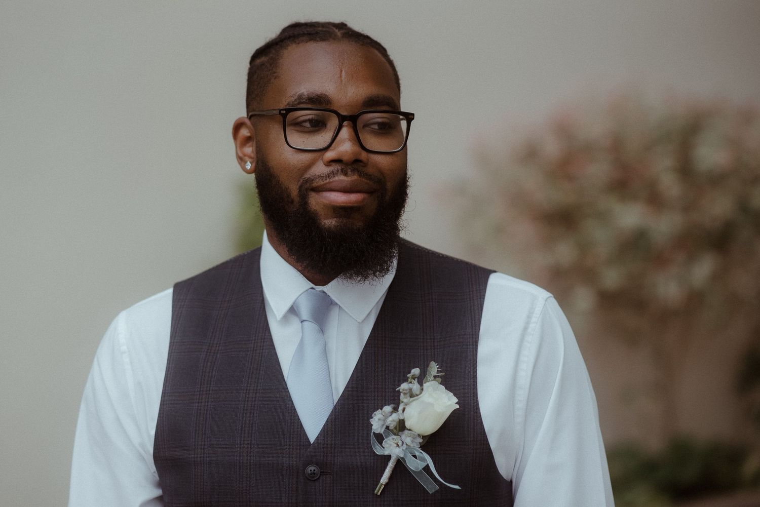 Groom in formal attire with glasses and boutonniere against neutral background.