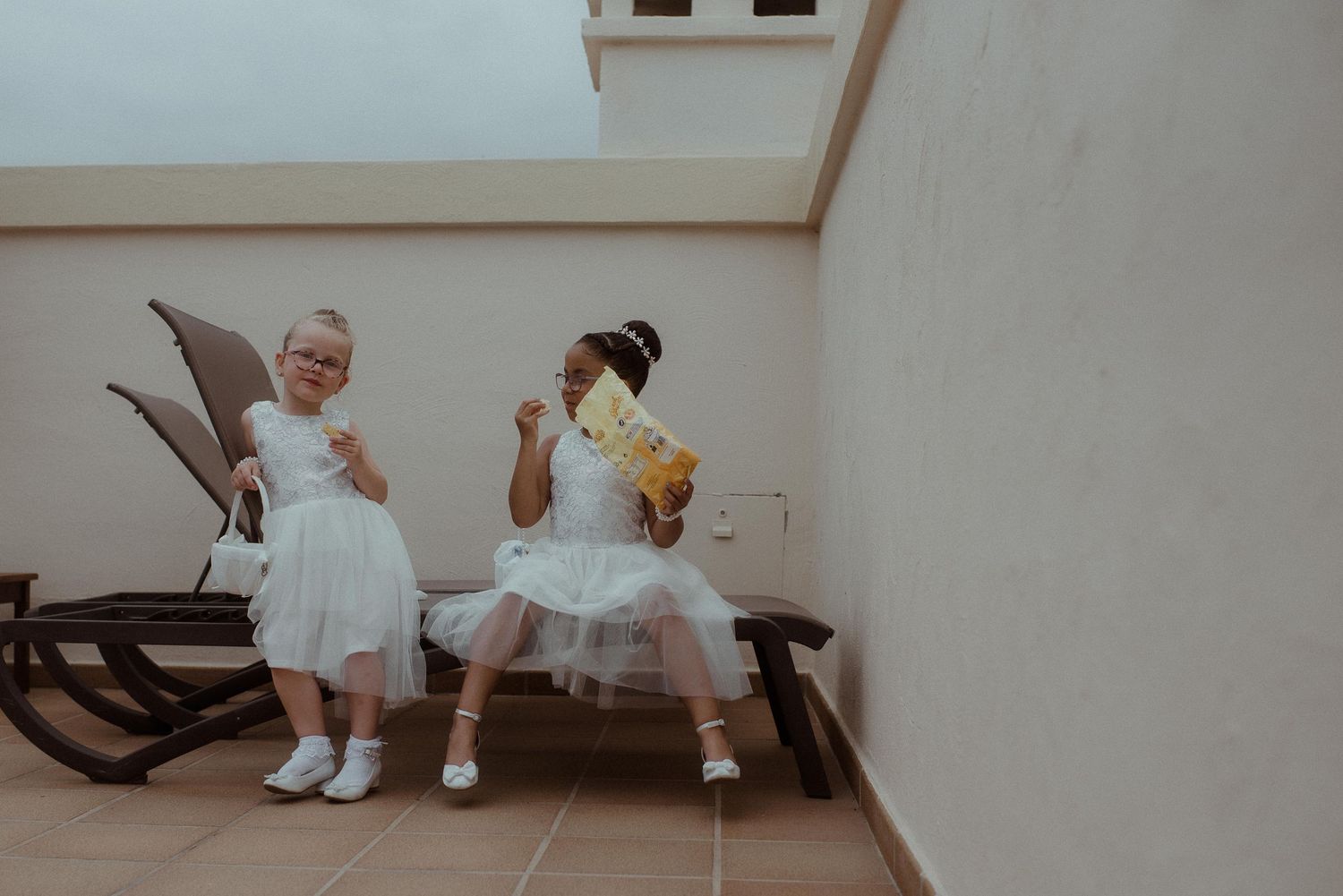 Two young flower girls in white dresses sitting on bench.