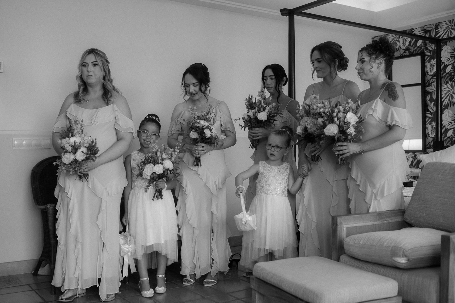 Black and white photo of bridesmaids and flower girls in flowing dresses holding bouquets lined up indoors.