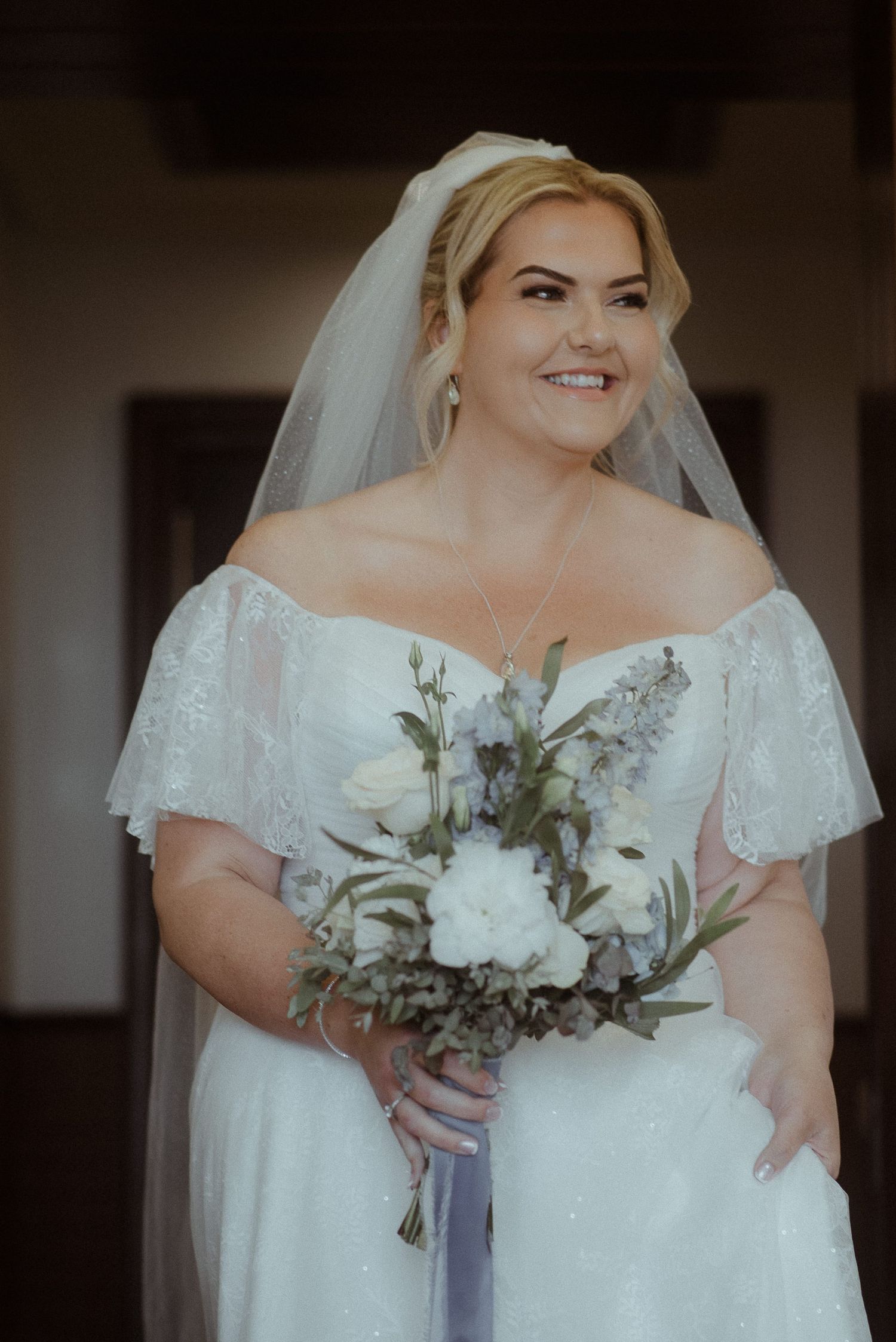 A bride in a sparkly off-shoulder gown smiles while holding a white and purple bouquet.
