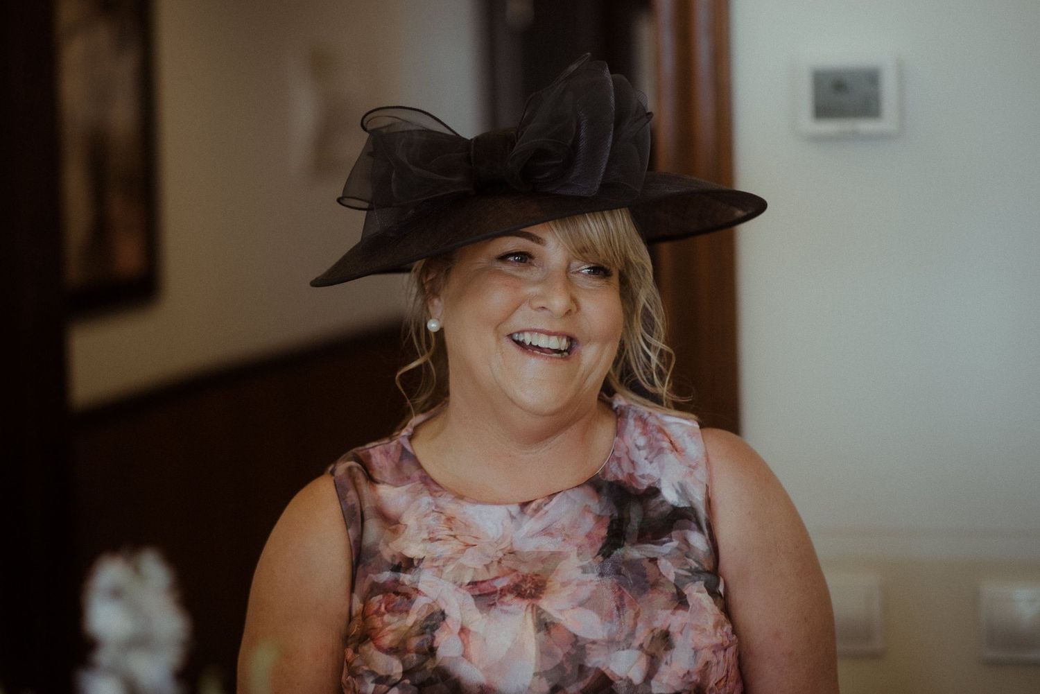 A wedding guest wearing a floral dress and large black hat smiles indoors.
