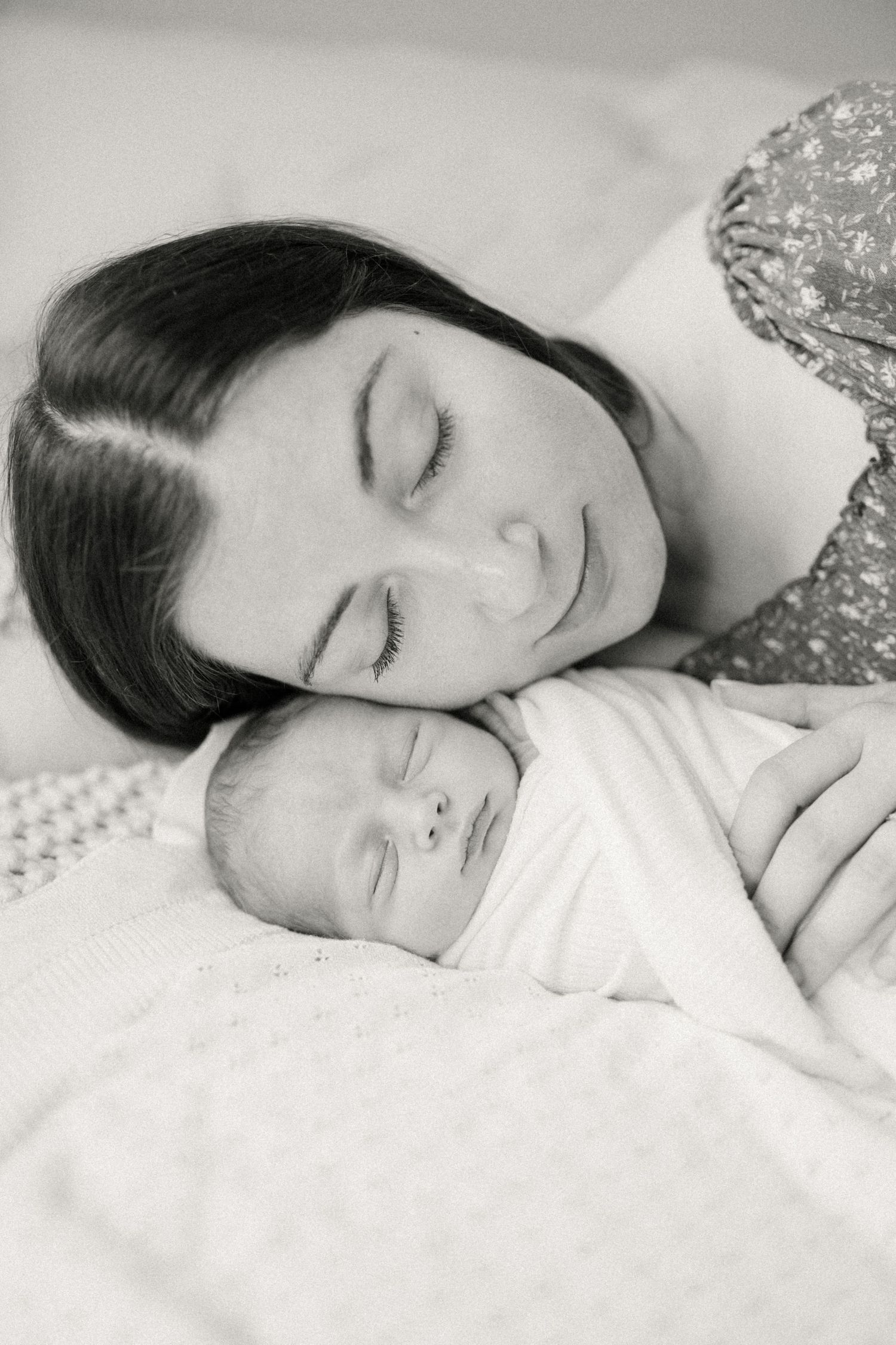 A tender black and white portrait of a parent and newborn baby sleeping peacefully together on white bedding.