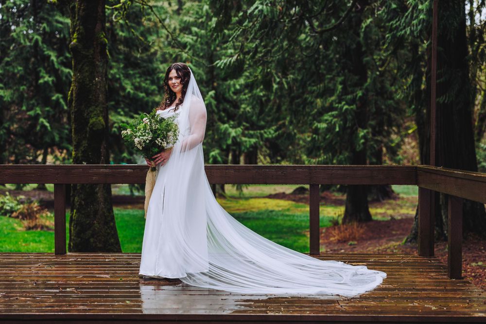 A white wedding gown with a flowing cathedral veil stands elegantly on a wooden bridge in a lush forest setting.