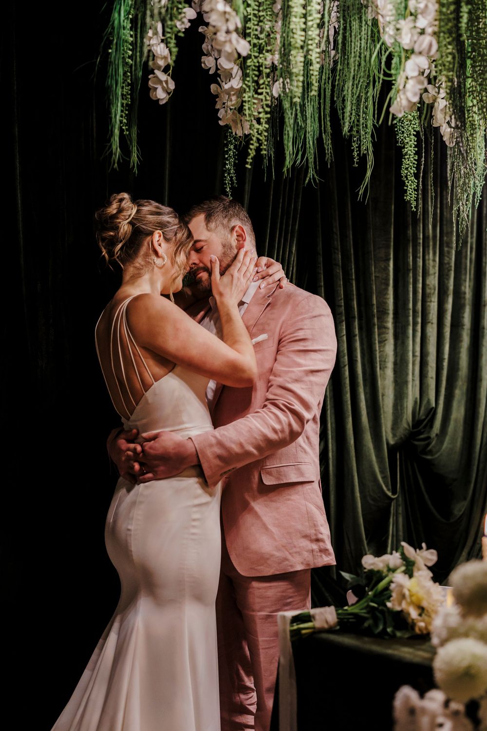 A romantic wedding couple embraces under hanging greenery and floral decorations at their intimate ceremony.