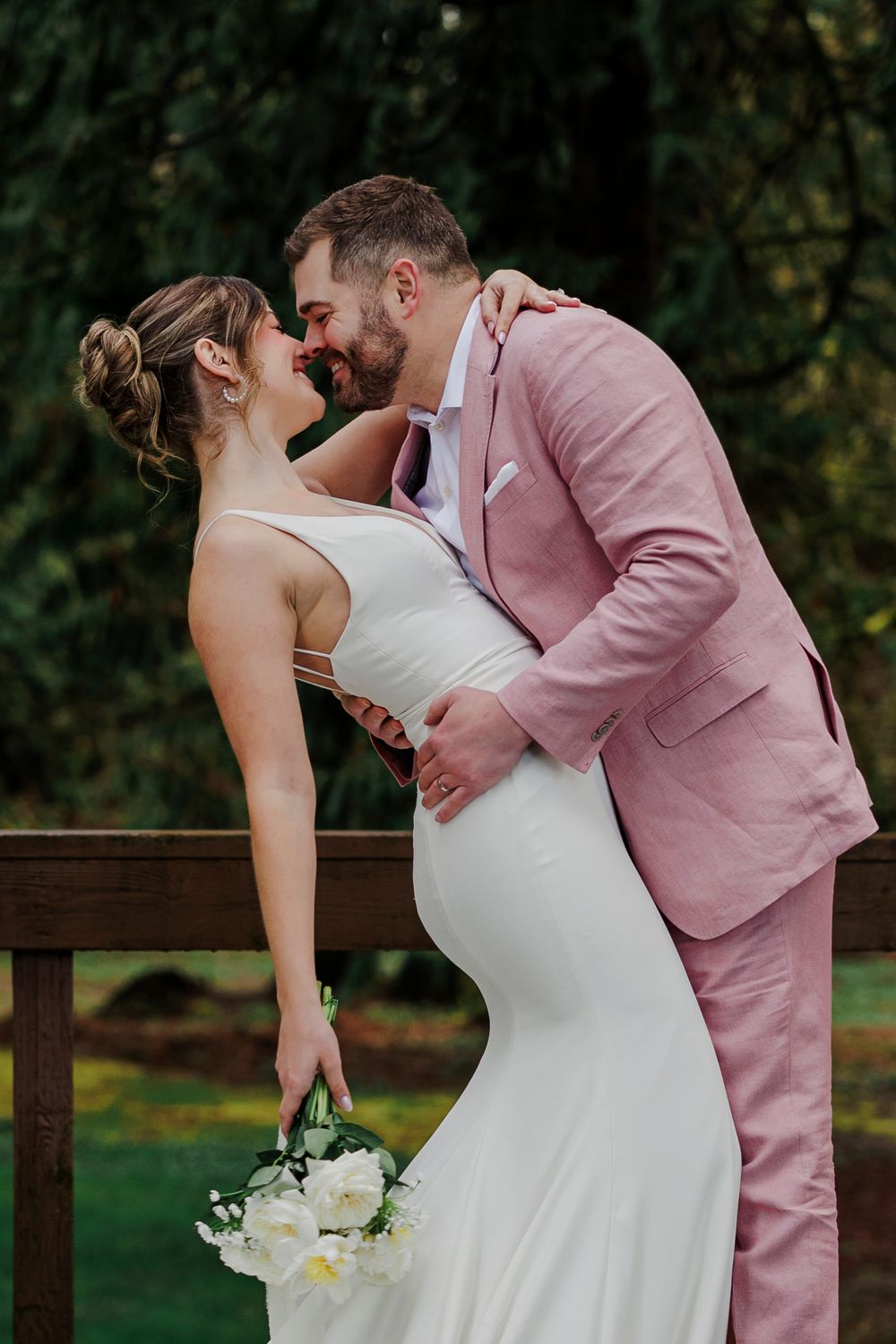 A romantic wedding photo showing a couple embracing outdoors, with the bride in white and groom in a pink suit.