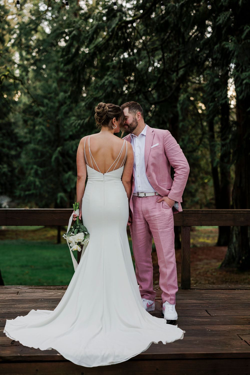 A wedding couple stands on a wooden deck, with the bride in white gown and groom wearing a pink suit in a forest setting.