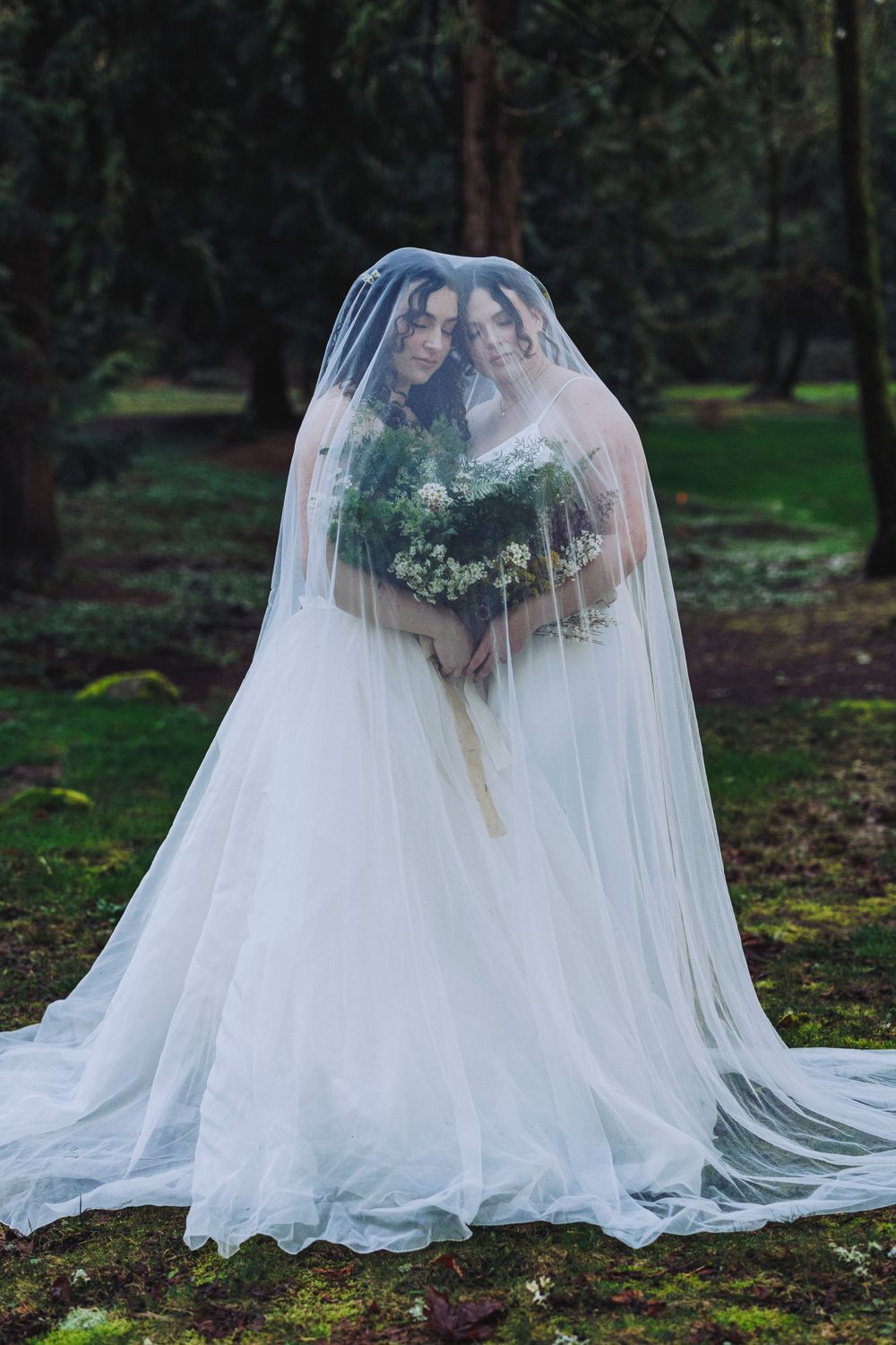Two brides share an intimate moment under a flowing white veil in a moody forest setting.